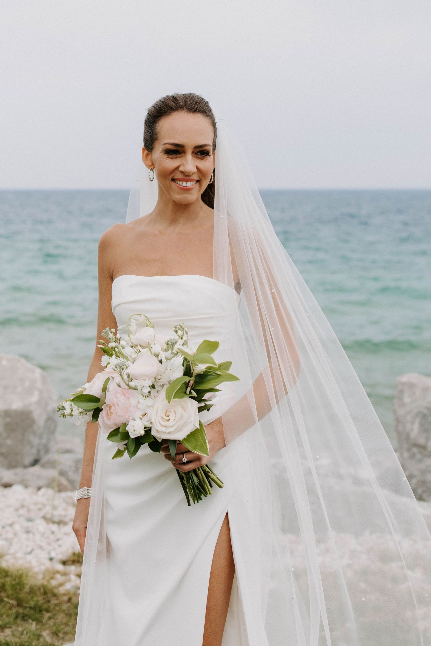 Elegant bride in strapless white gown with slit holding bouquet by ocean.