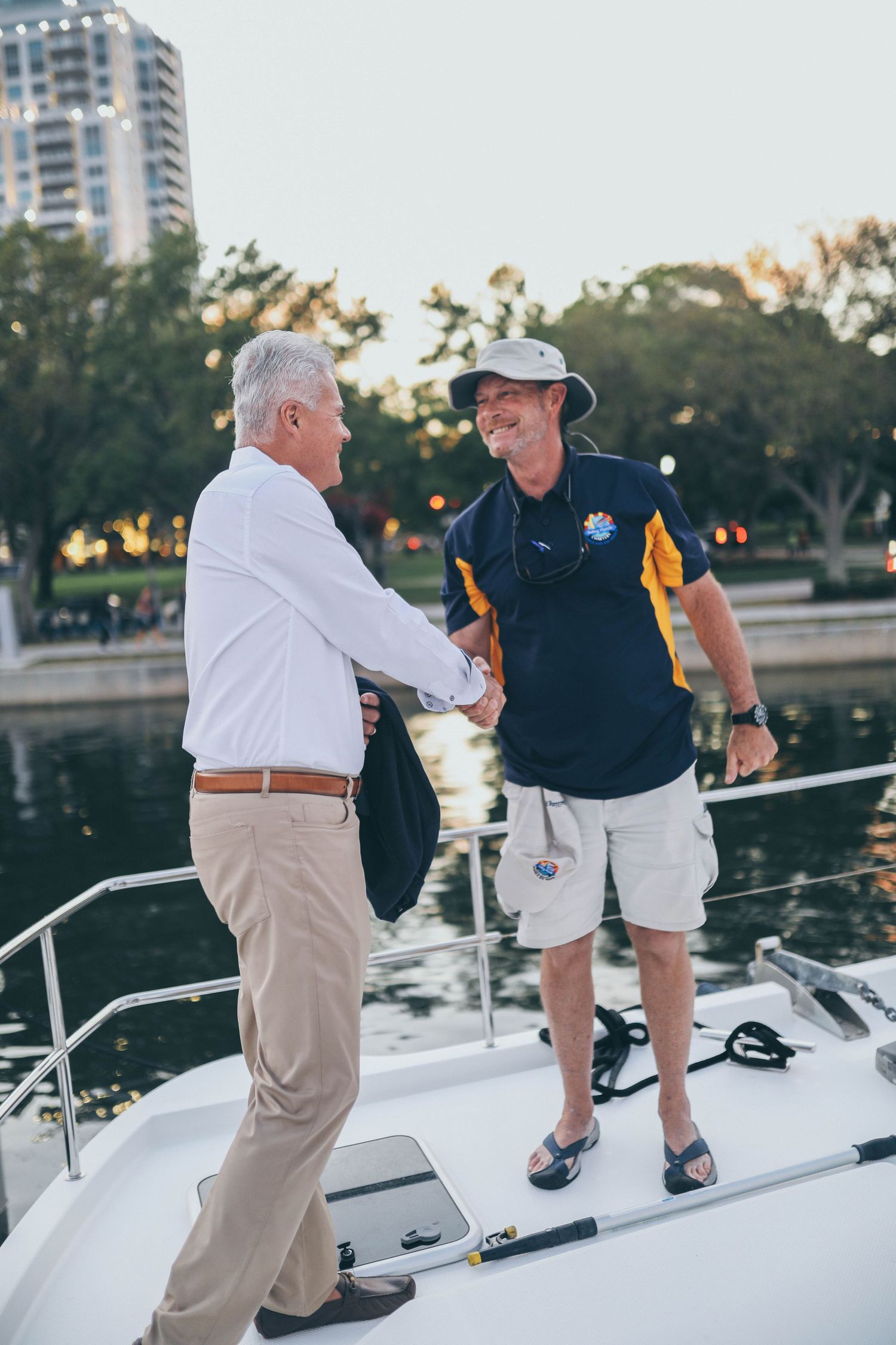 Two men shake hands while standing on the deck of a boat at sunset with a high-rise building visible in background.