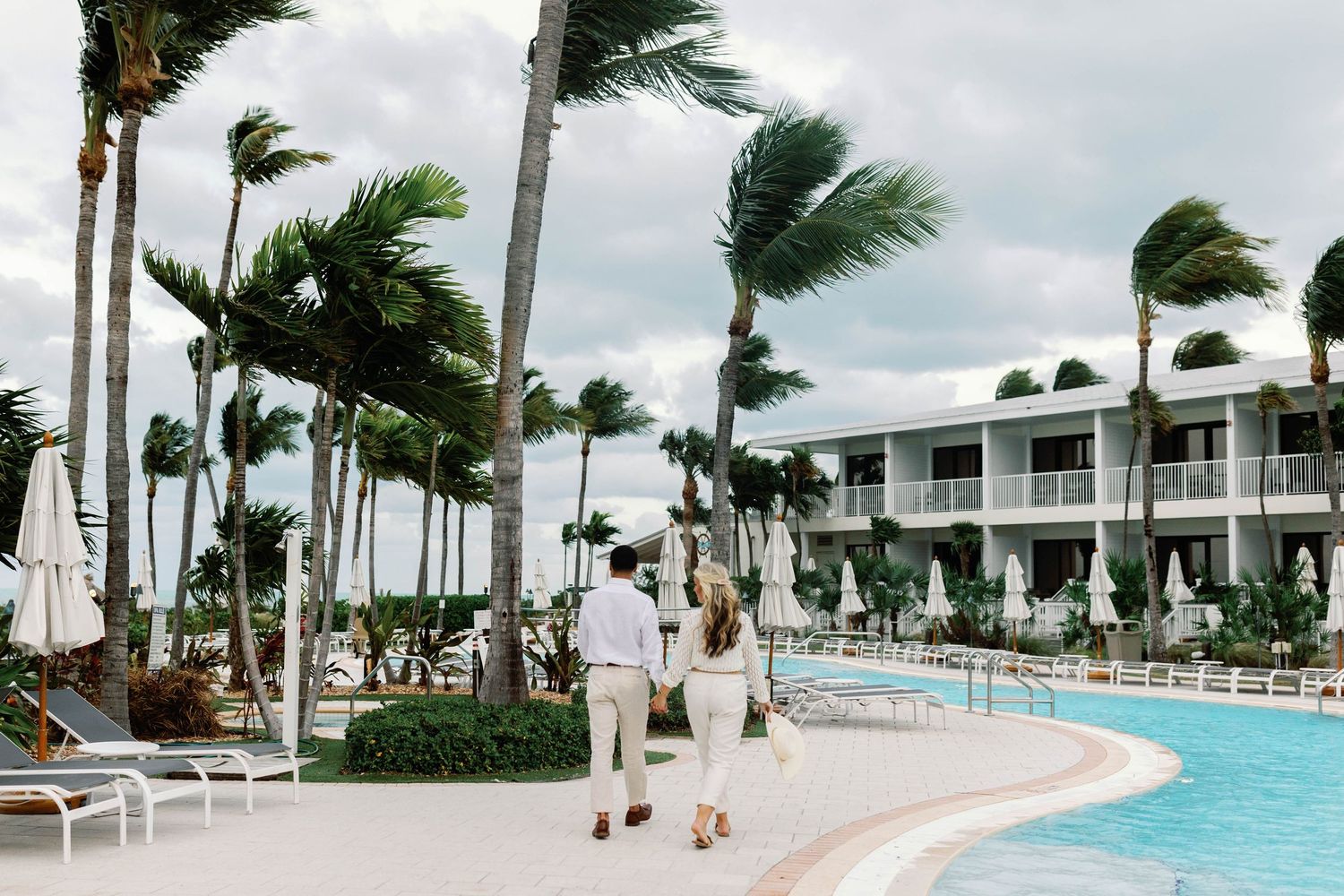 Palm trees sway in the wind along a luxury resort pool deck with white lounge chairs and turquoise water.