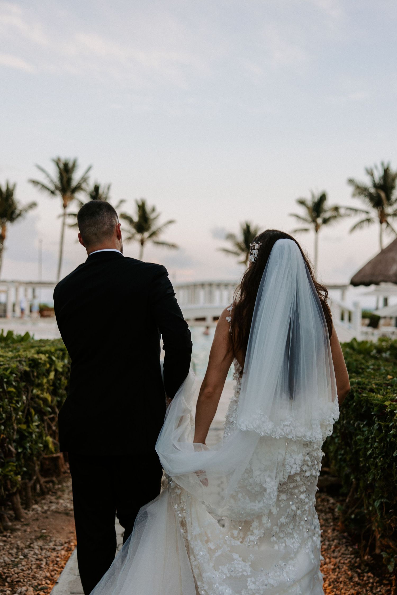 Couple walking along tropical beach path with flowing wedding veil.