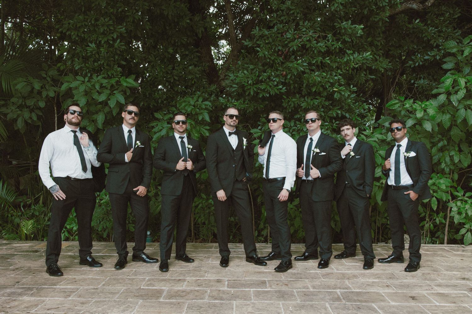 Group of groomsmen in black suits doing coordinated jump photo against garden backdrop.