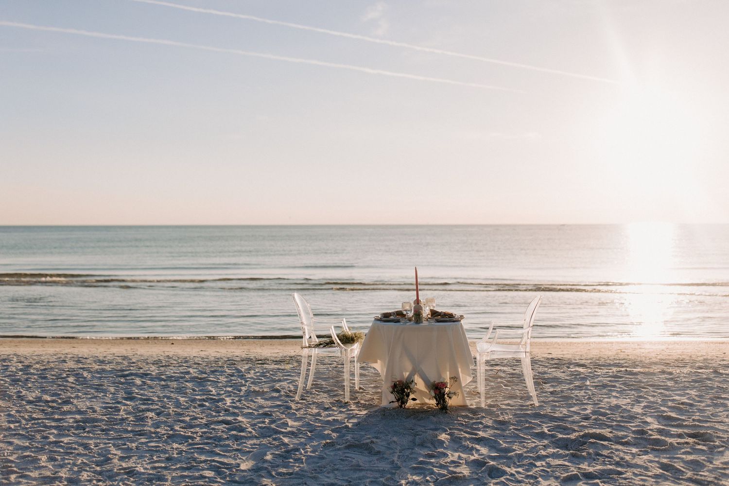 Romantic beachside dining setup with white table and chairs on sandy shore during golden sunset over ocean waves.
