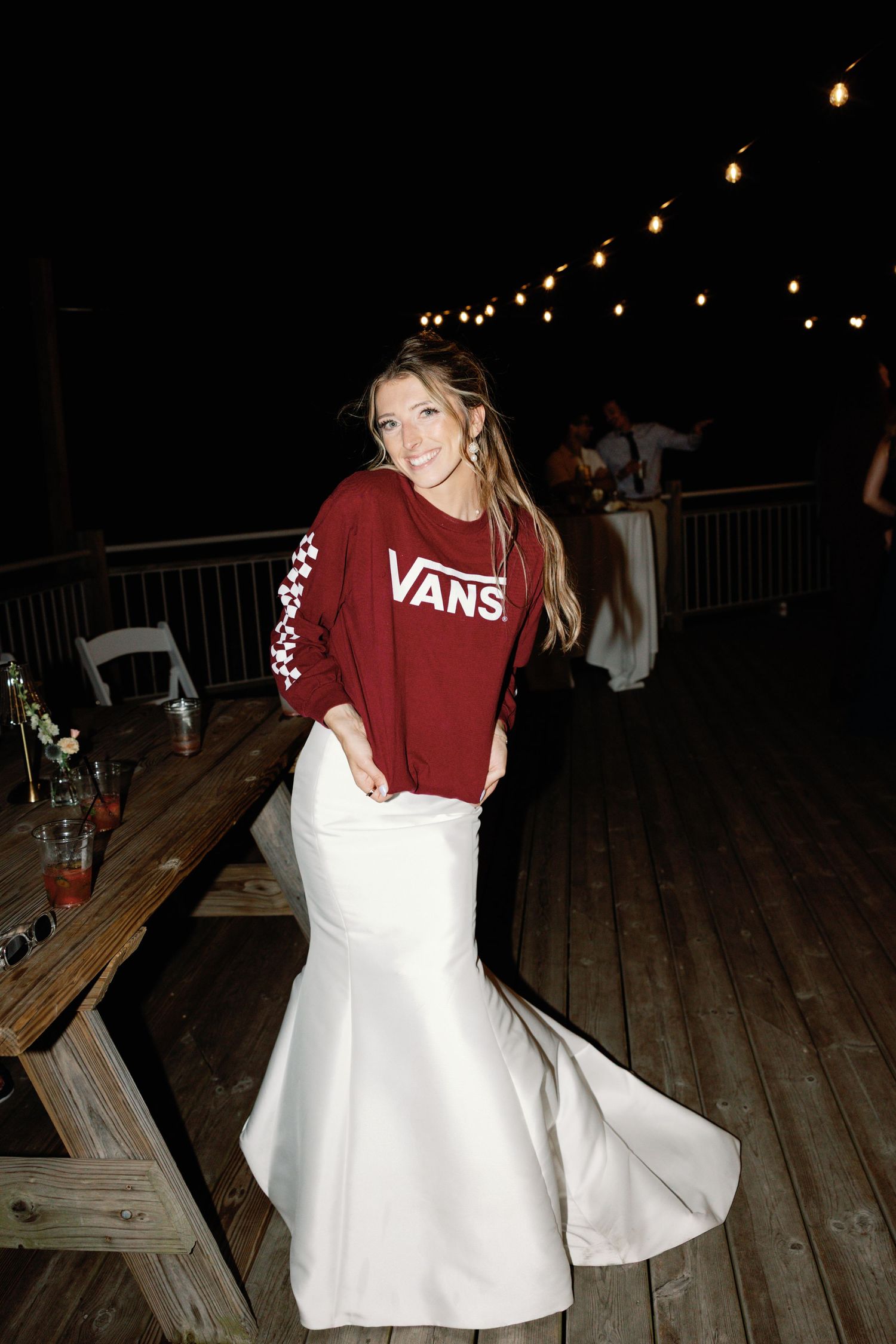 A person wearing a red Vans sweatshirt and white dress dances at a nighttime outdoor event with string lights.