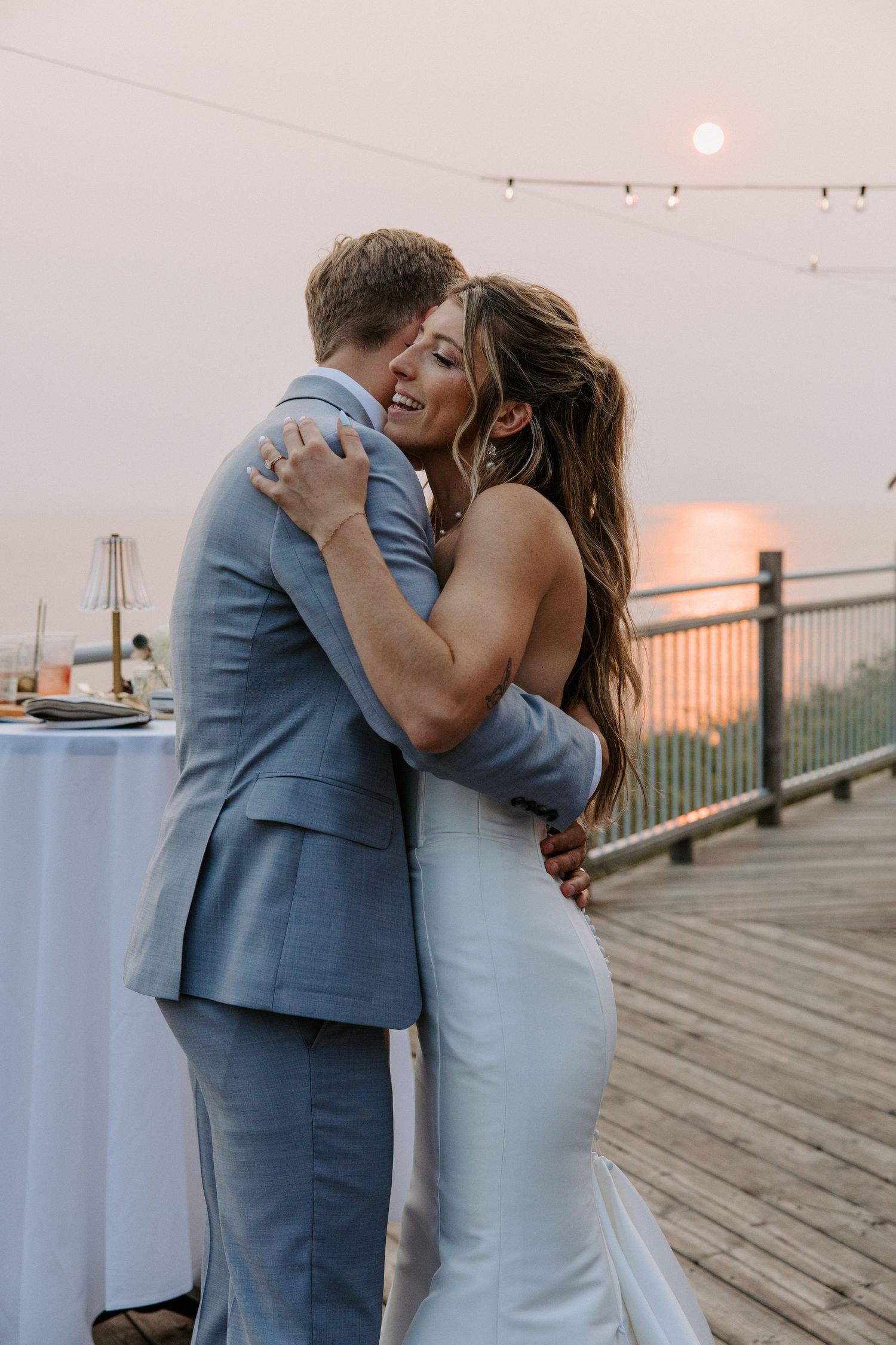 Newlyweds share an intimate moment on a wooden deck at dusk with string lights in the background.