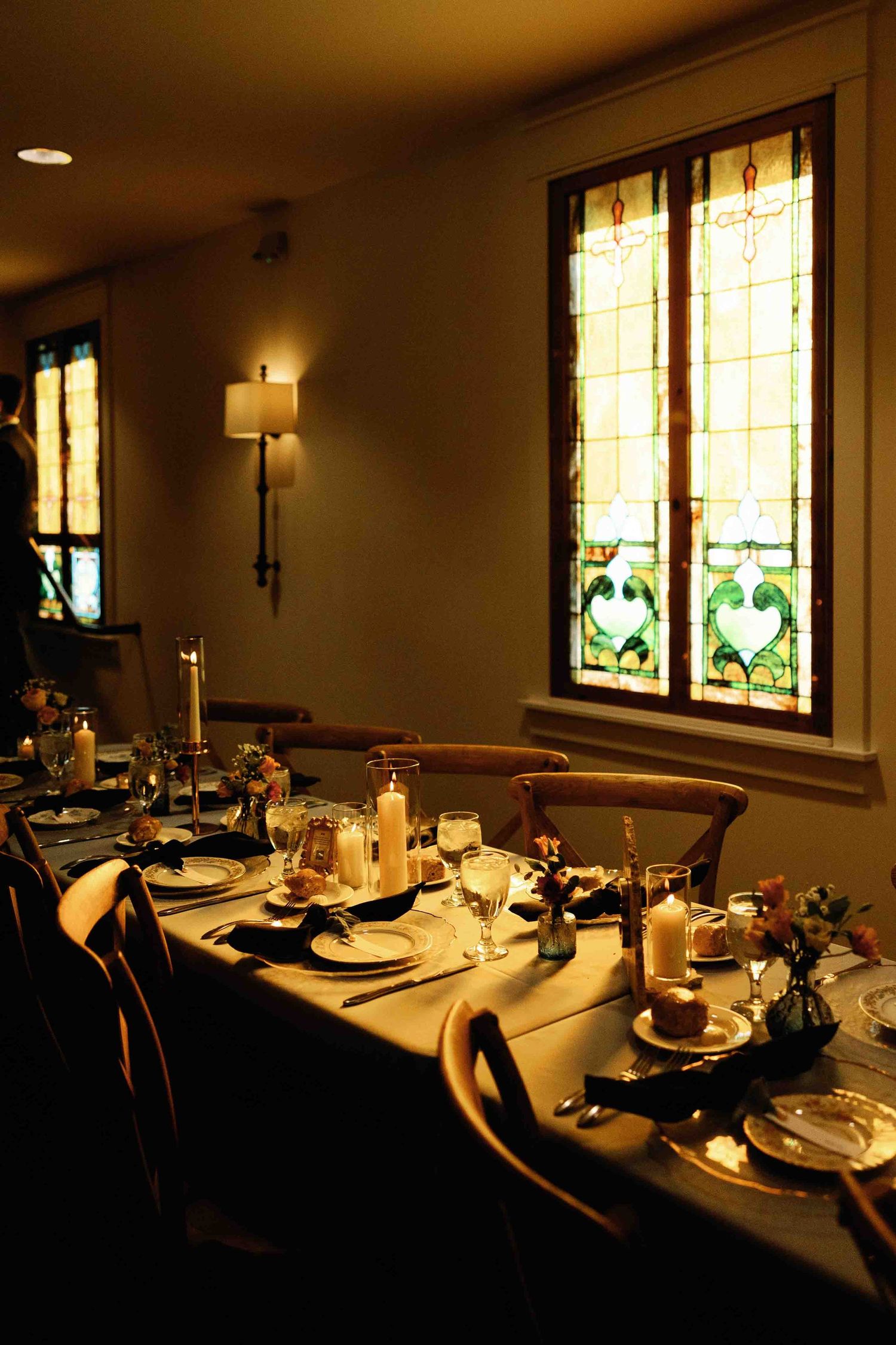 Wedding reception dining room with stained glass windows and elegant table settings in warm lighting.