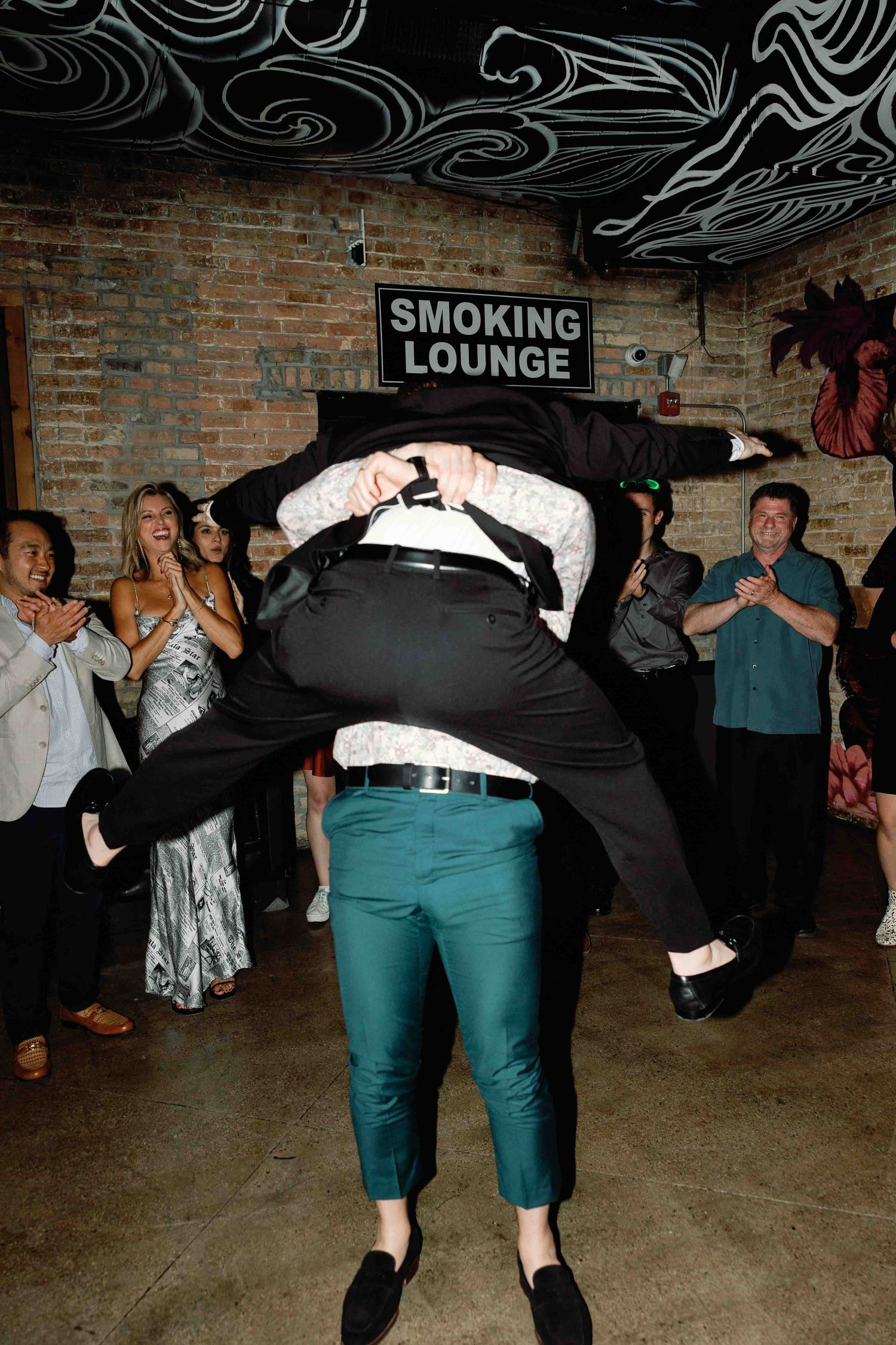 Partygoers perform dance moves in a basement lounge with exposed brick walls.