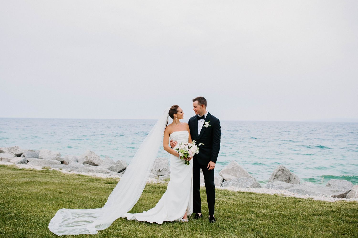 Coastal wedding scene with couple and flowing veil against ocean landscape.