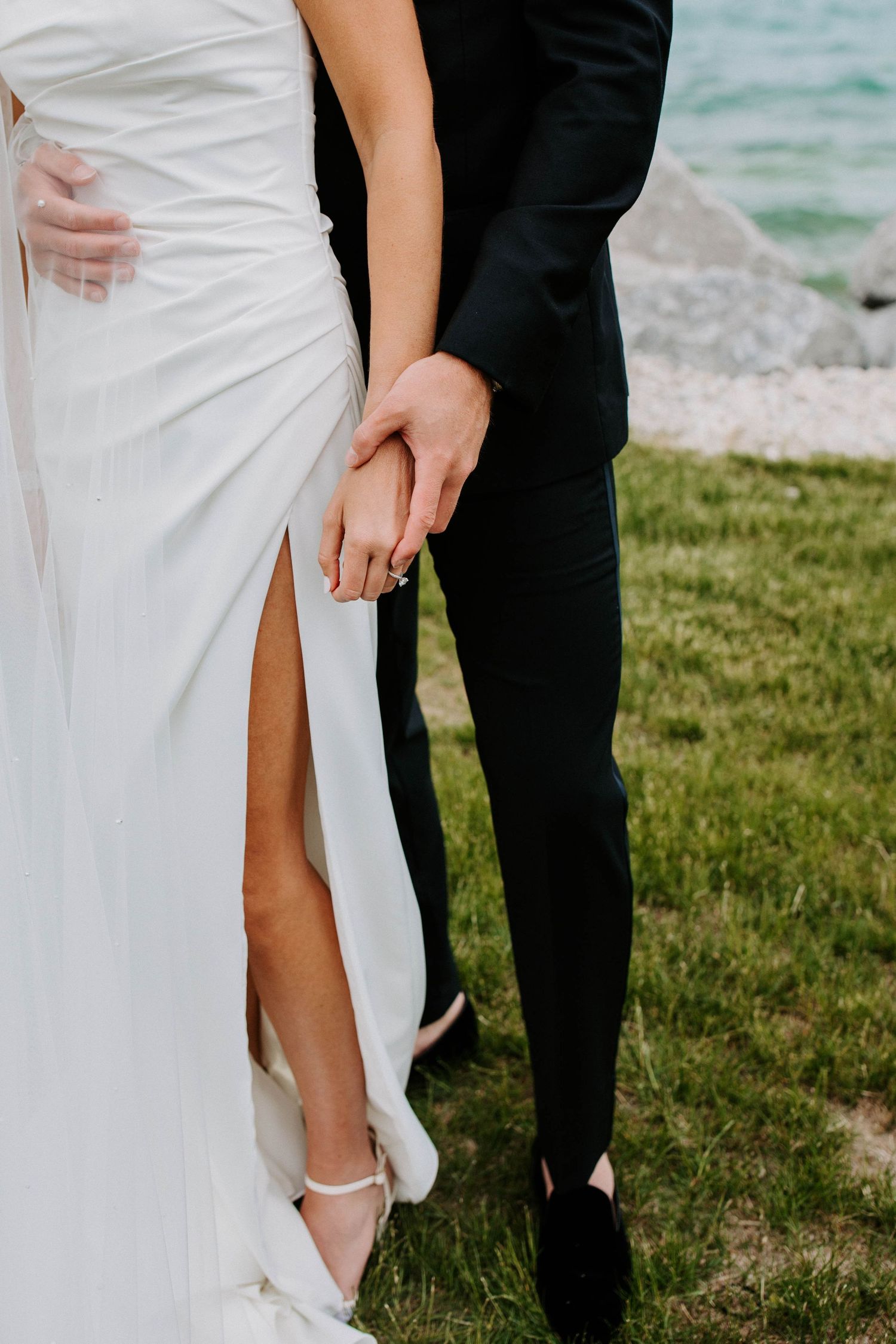 Close-up of bride and groom holding hands, showing white wedding dress detail and black tuxedo.