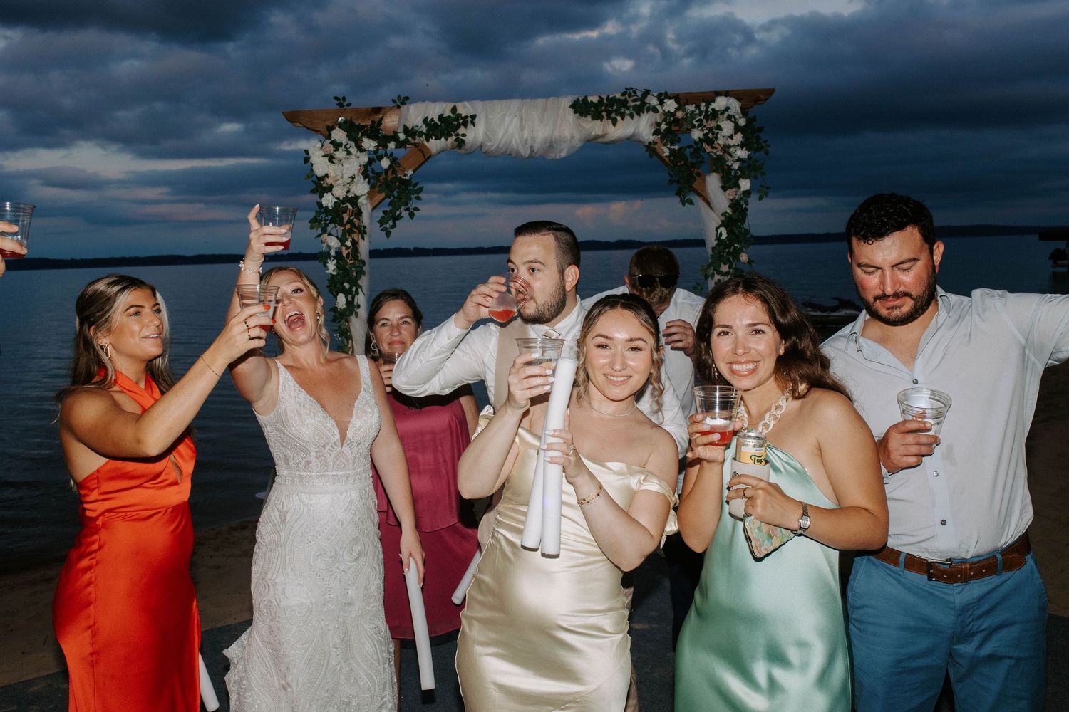 Wedding guests raise champagne glasses in a celebratory toast at a beachside reception under dramatic clouds.