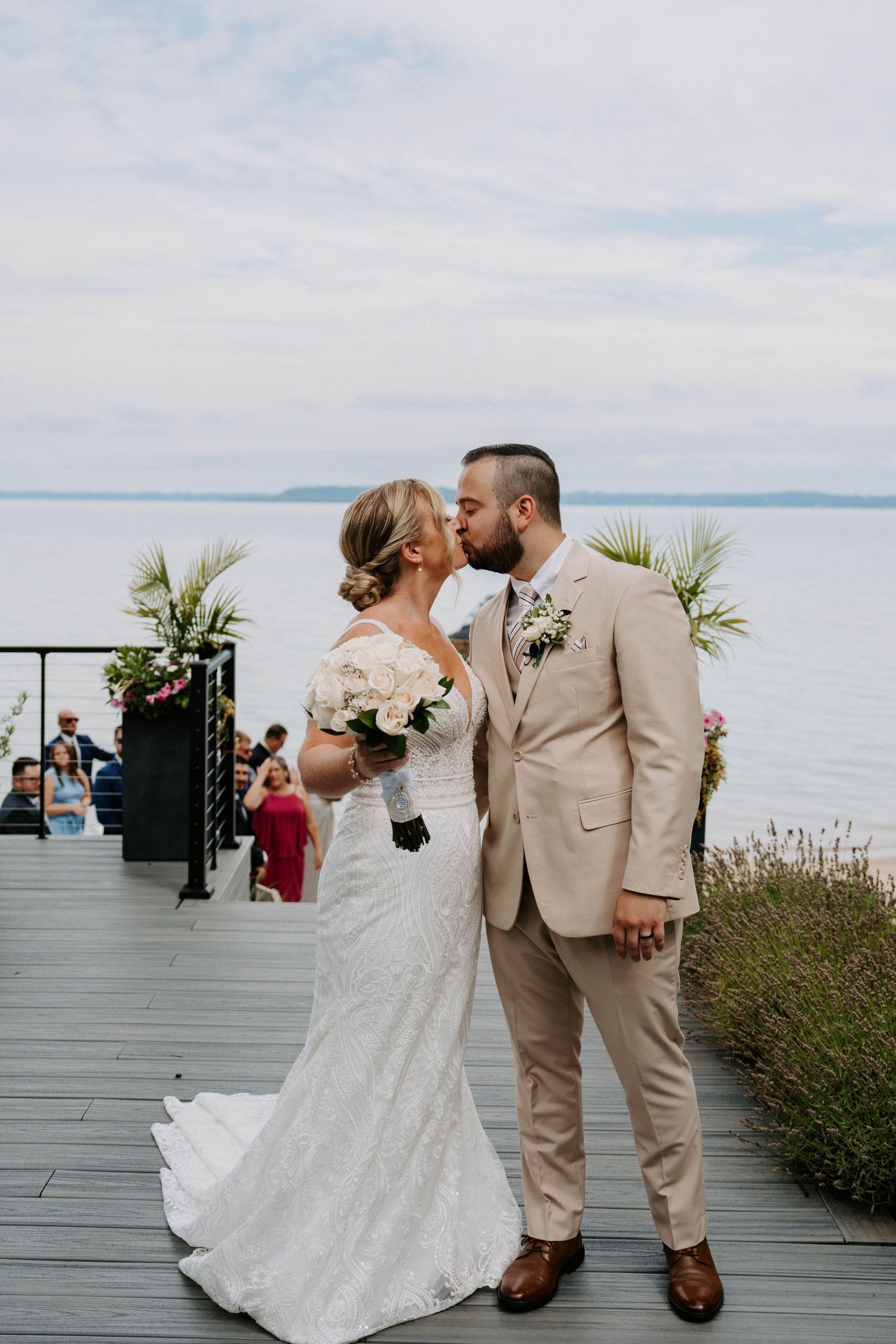 Newlyweds share their first kiss on a waterfront deck decorated with tropical plants.