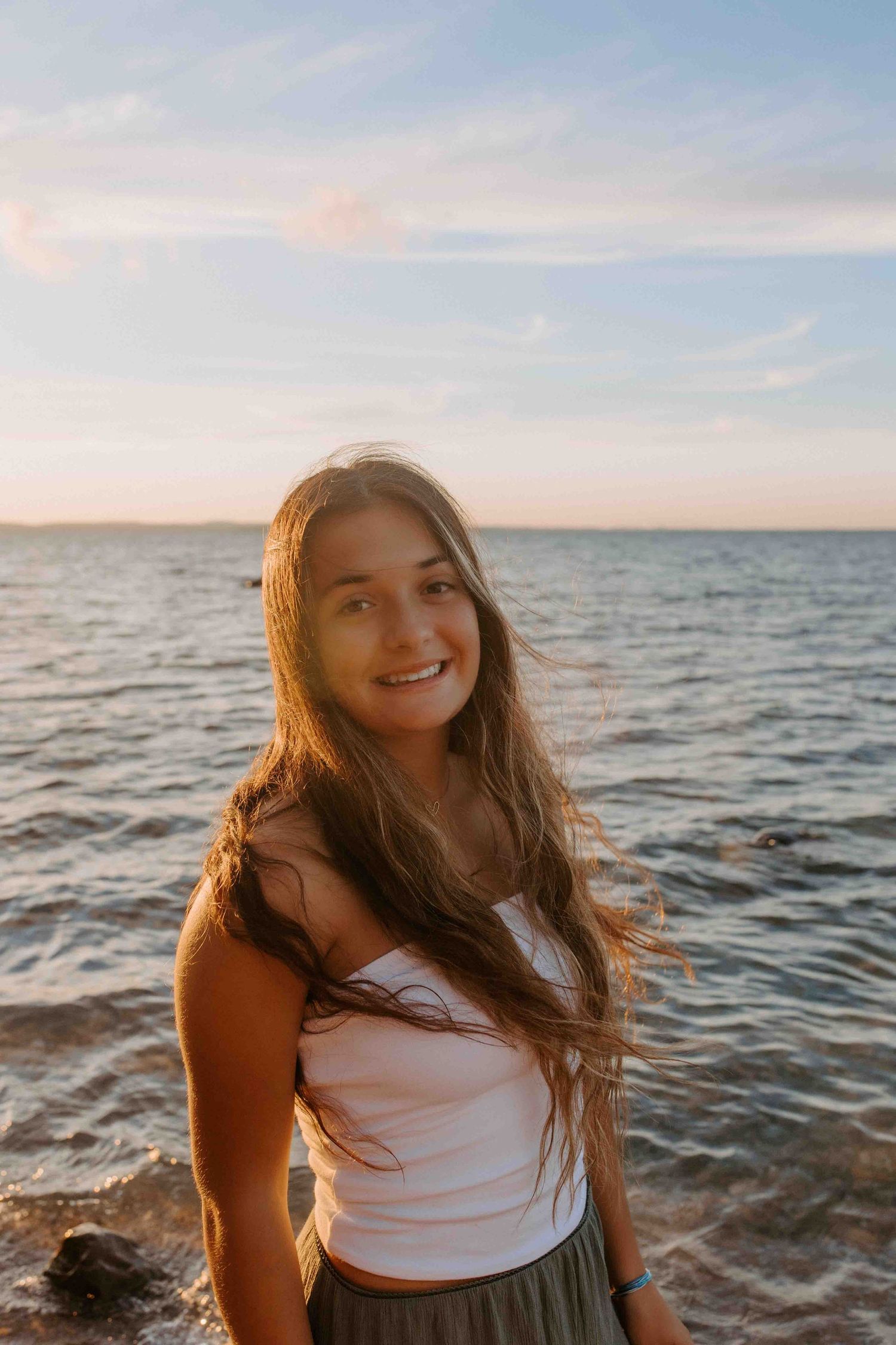 A person in a white tank top poses by the ocean during sunset with long hair flowing in the breeze.