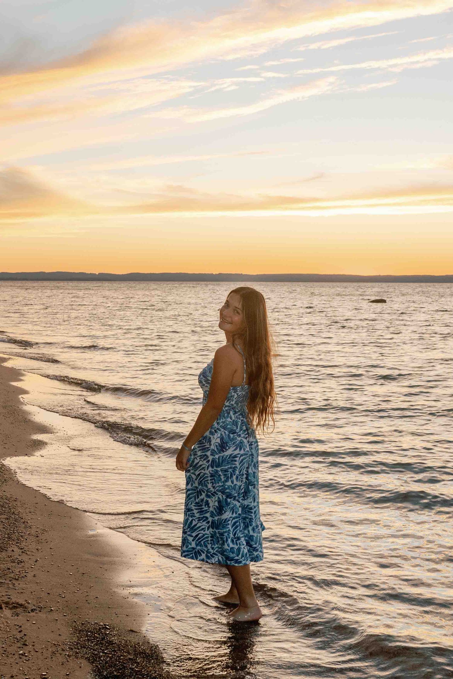 Waves crash onto the shore as someone in a blue dress enjoys the warm sunset light.