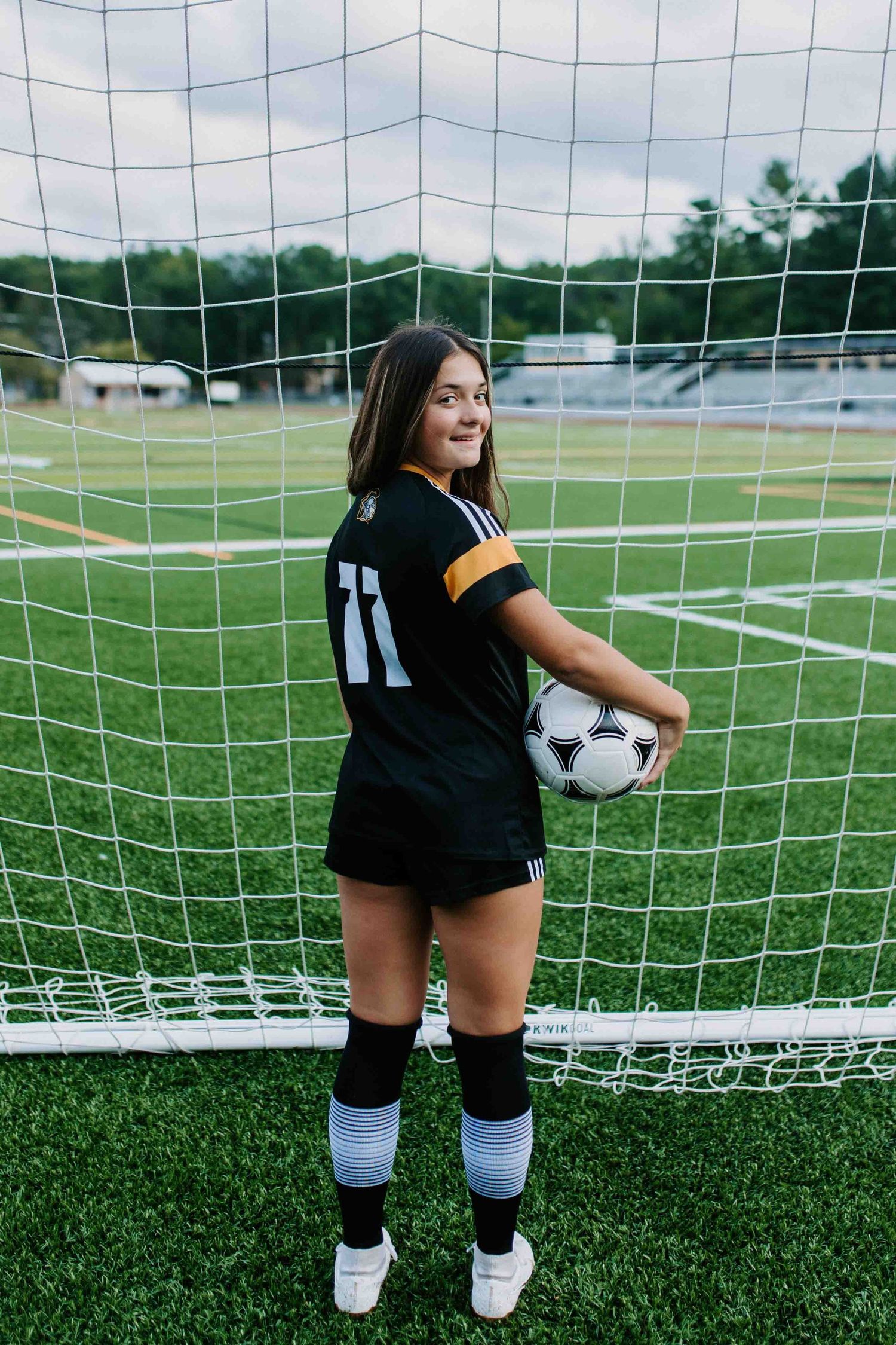 Soccer player in black uniform holding ball while posing by goal net on green turf field.