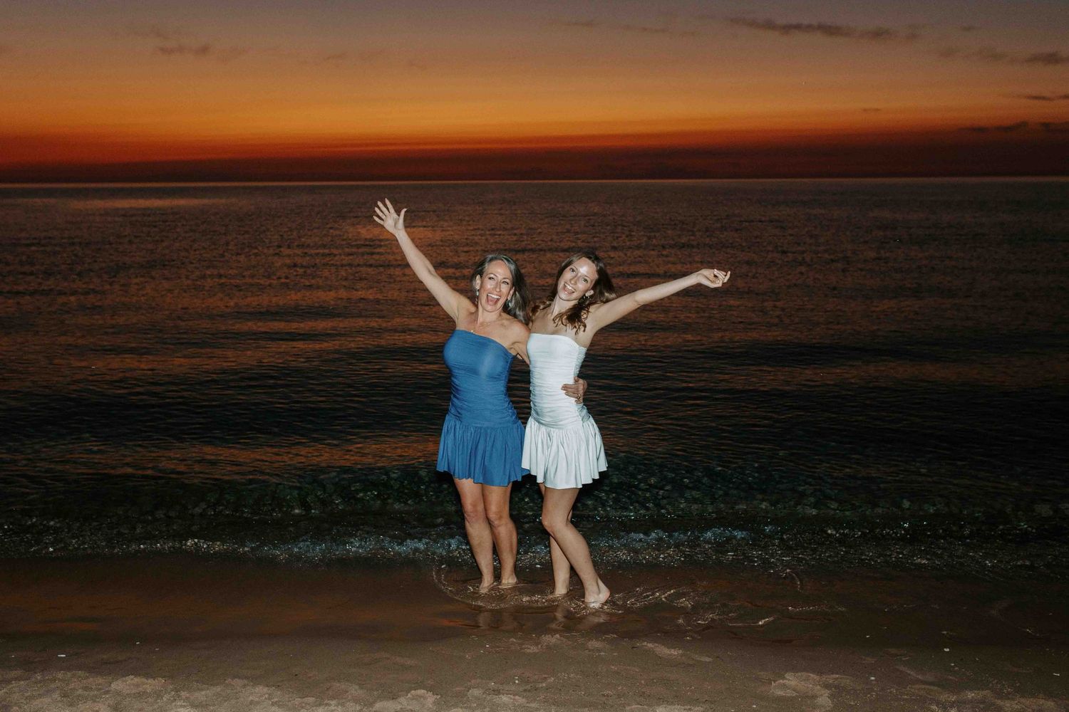 Two people dance joyfully on the beach at sunset with the ocean waves in the background.