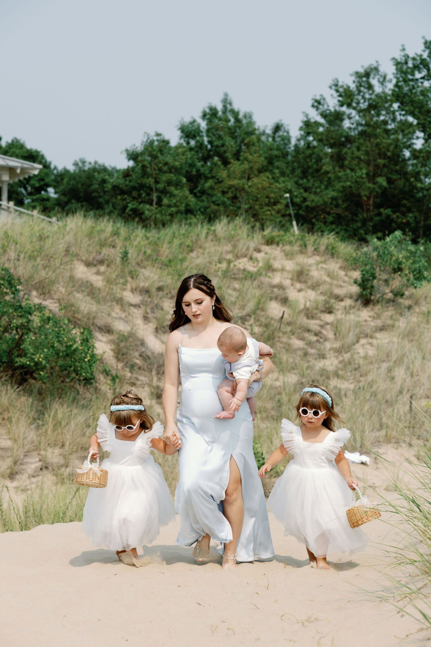Bridesmaids in white dresses sitting together on a sandy beach dune.