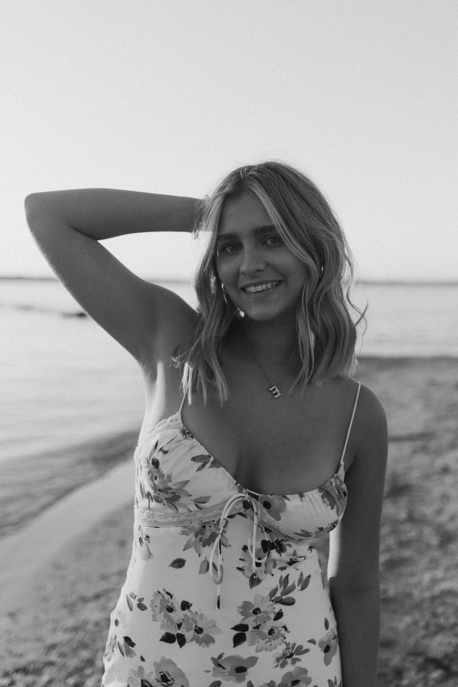 Black and white portrait of woman in floral dress posing on beach at sunset.
