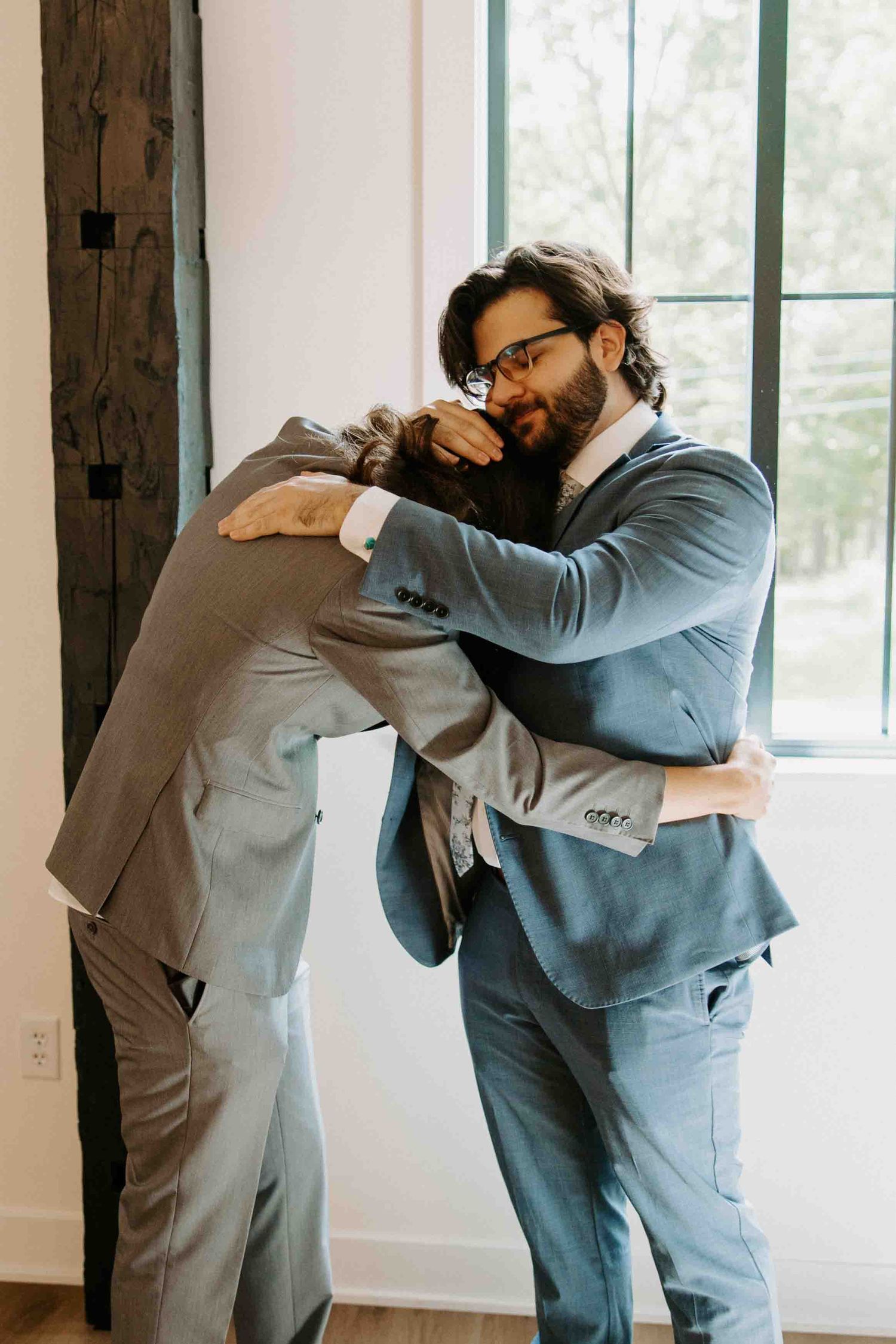 Two people in gray suits embracing in a bright room with windows.