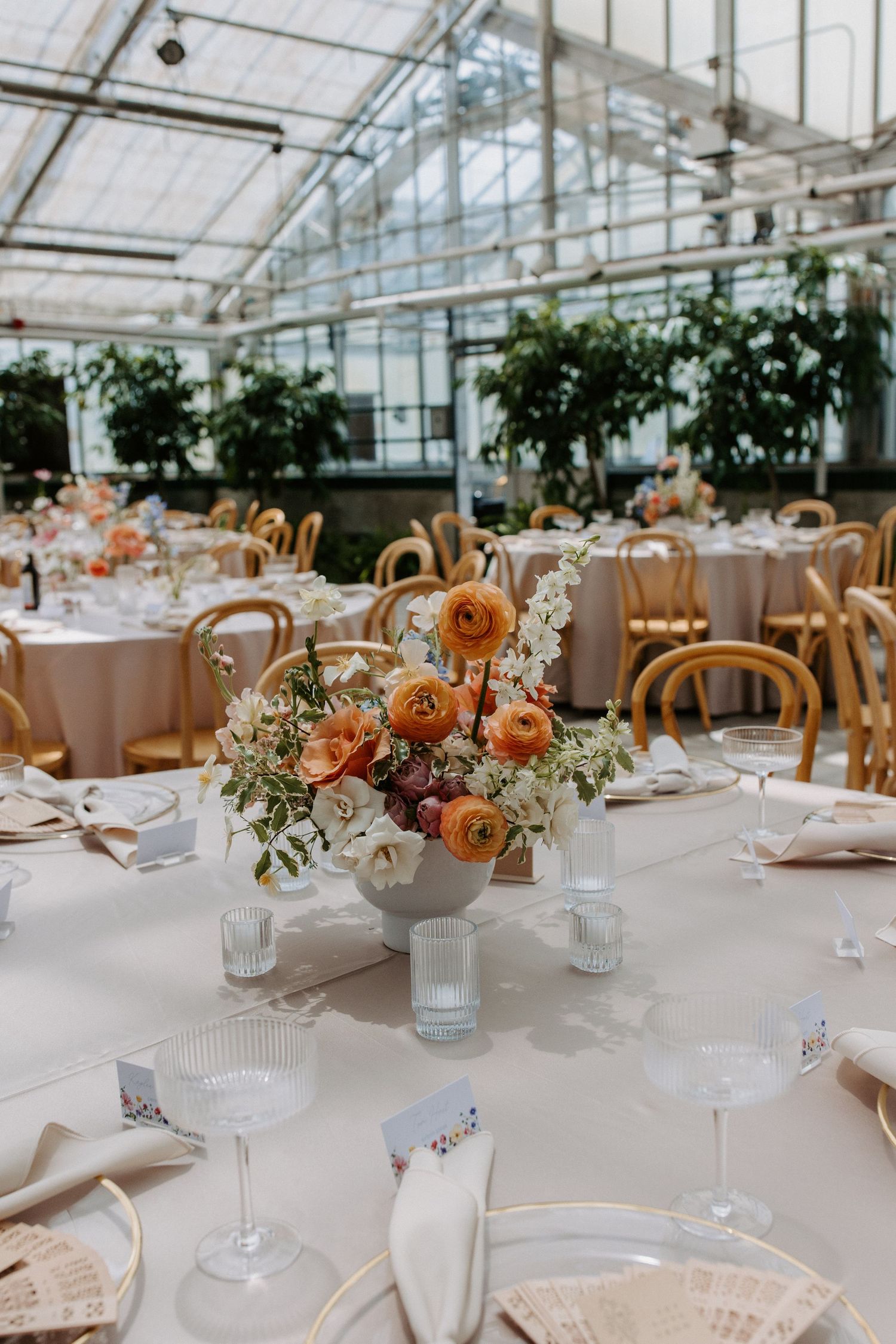 Elegant greenhouse reception setup with round tables, floral centerpieces and bentwood chairs.