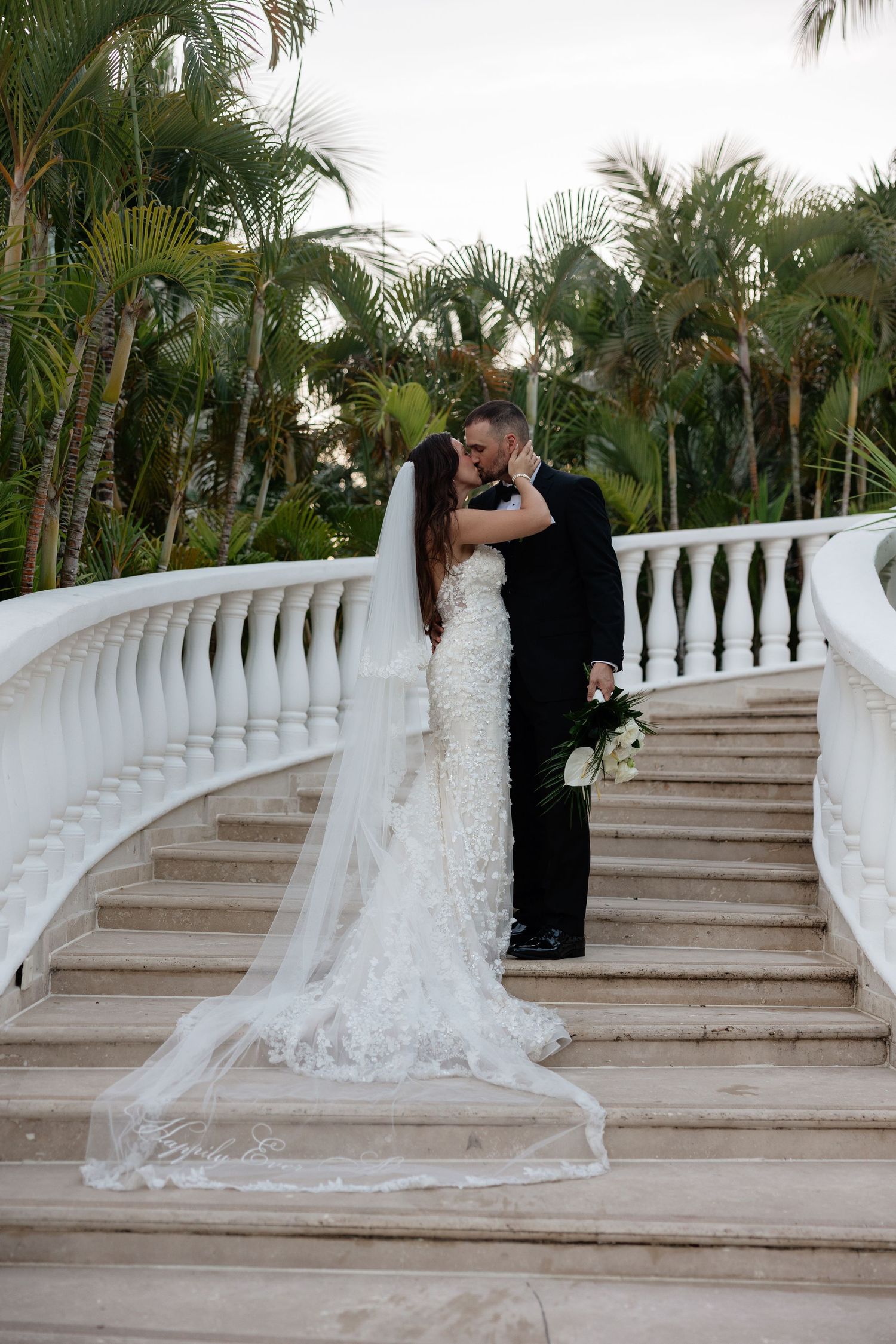 A romantic wedding couple embraces on an elegant white staircase surrounded by tropical palm trees in a garden setting at Dreams Tulum