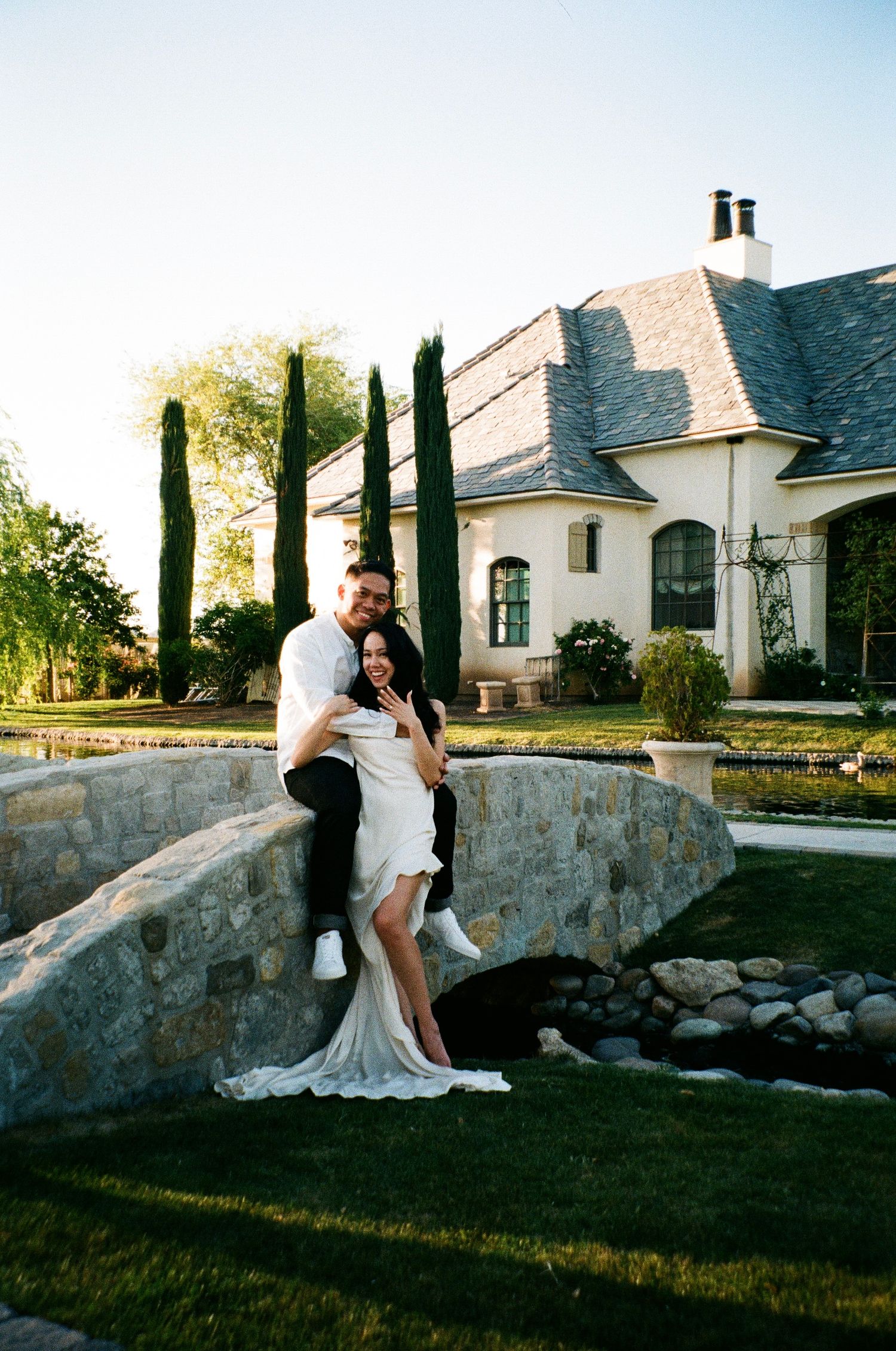 Romantic couple shares intimate moments on a stone wall outside an elegant white house with cypress trees at sunset.