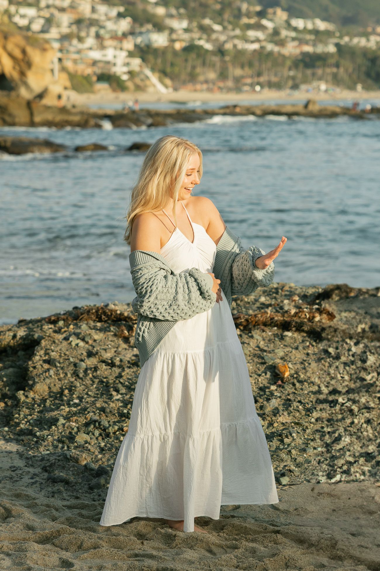 Woman in flowing white dress and gray sweater stands on rocky coastal beach at sunset with ocean waves behind.