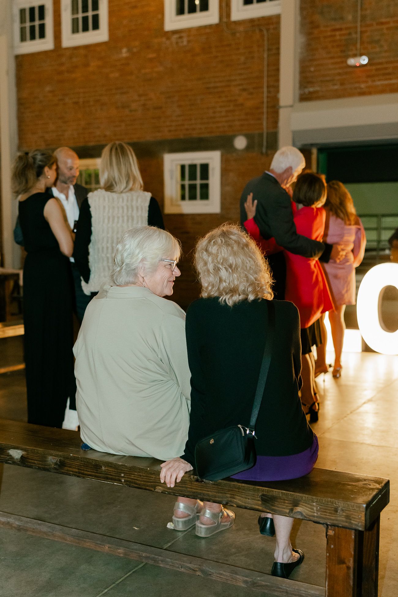 Wedding reception guests sit and mingle near illuminated decor in a rustic brick venue space.