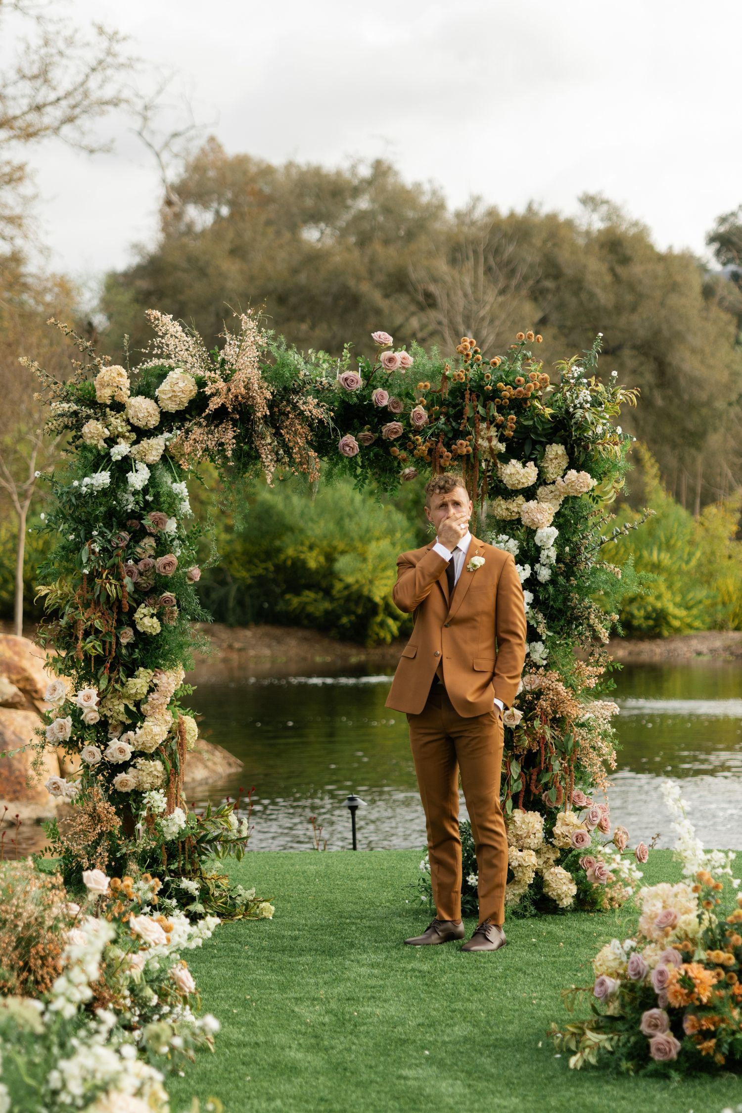 A floral wedding arch with white and peach blooms frames a scenic lakeside ceremony setting with green grass.