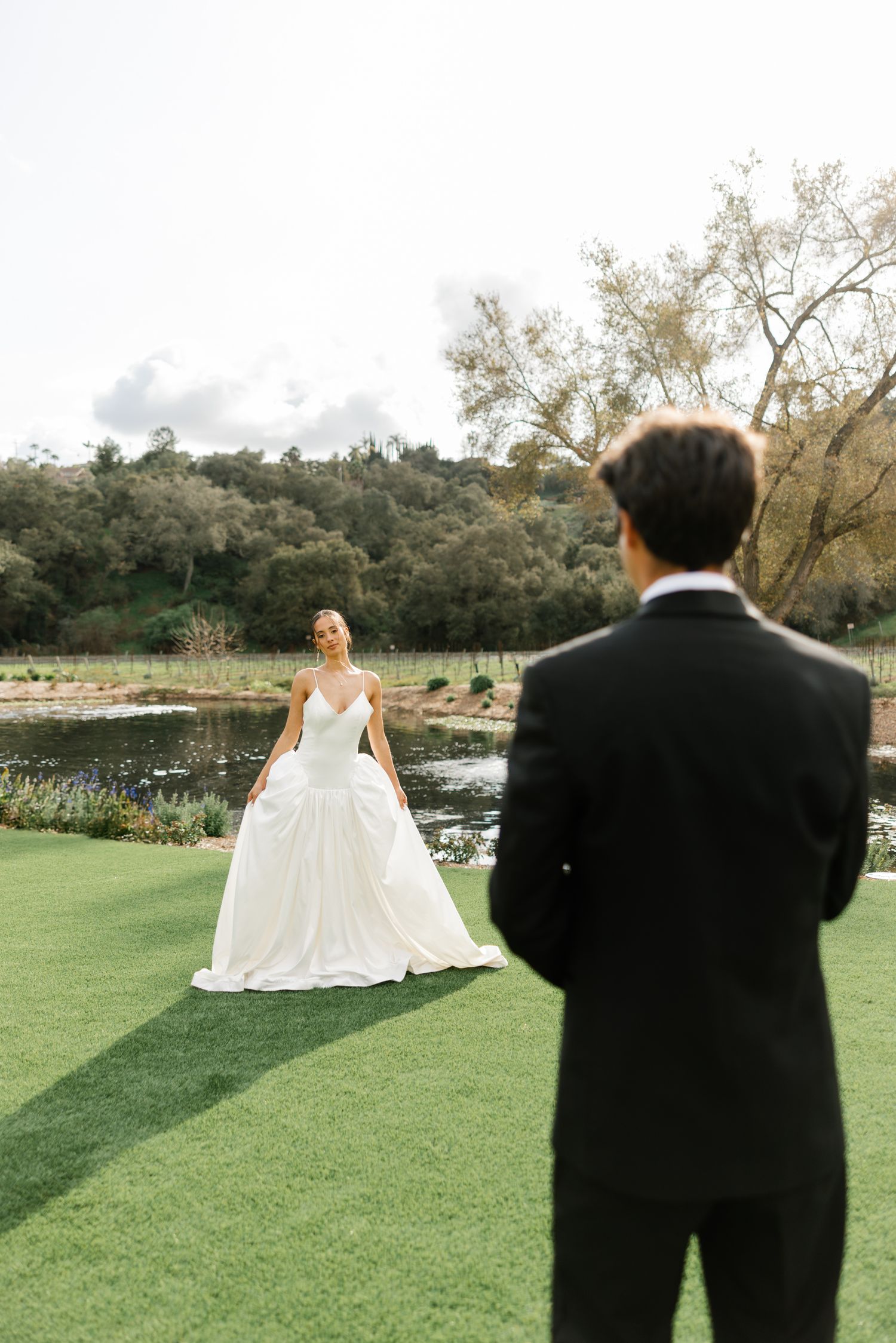 A couple shares a romantic moment on manicured lawn by a pond during their wedding photoshoot.