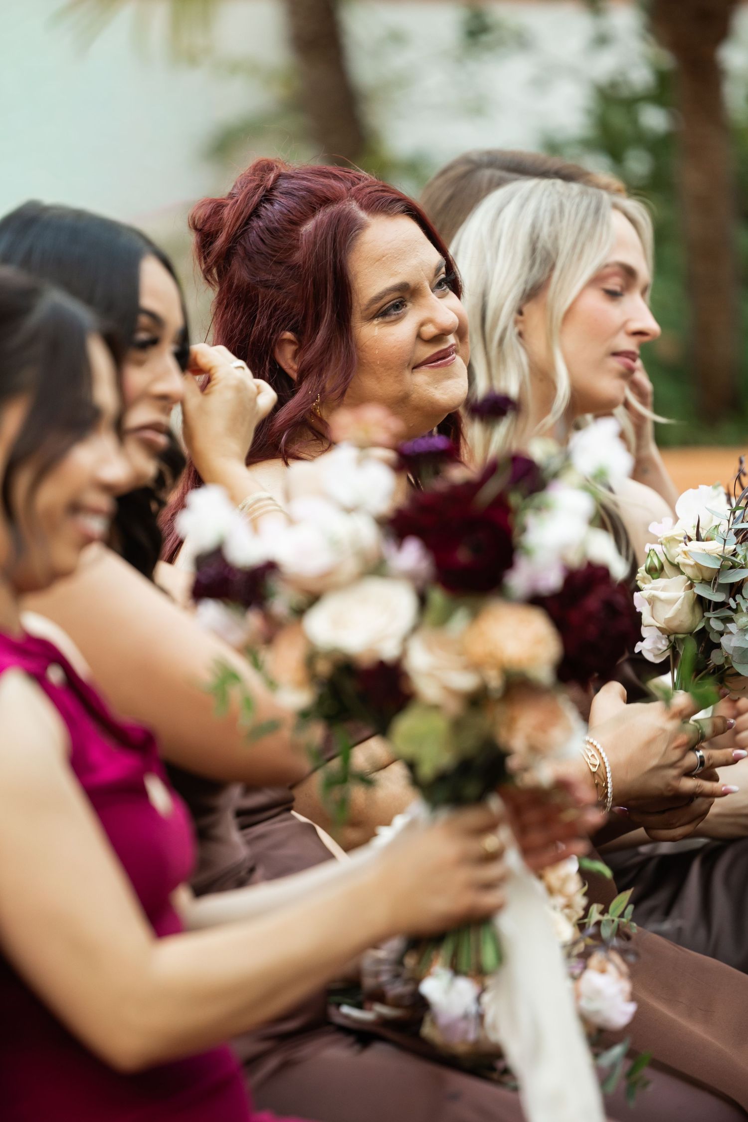 A line of bridesmaids in burgundy dresses hold white floral bouquets while laughing joyfully at an outdoor wedding.