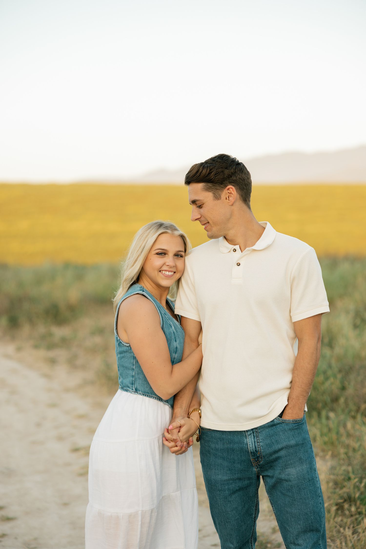 Couple in casual attire walking together on a dirt path through a sun-drenched field.