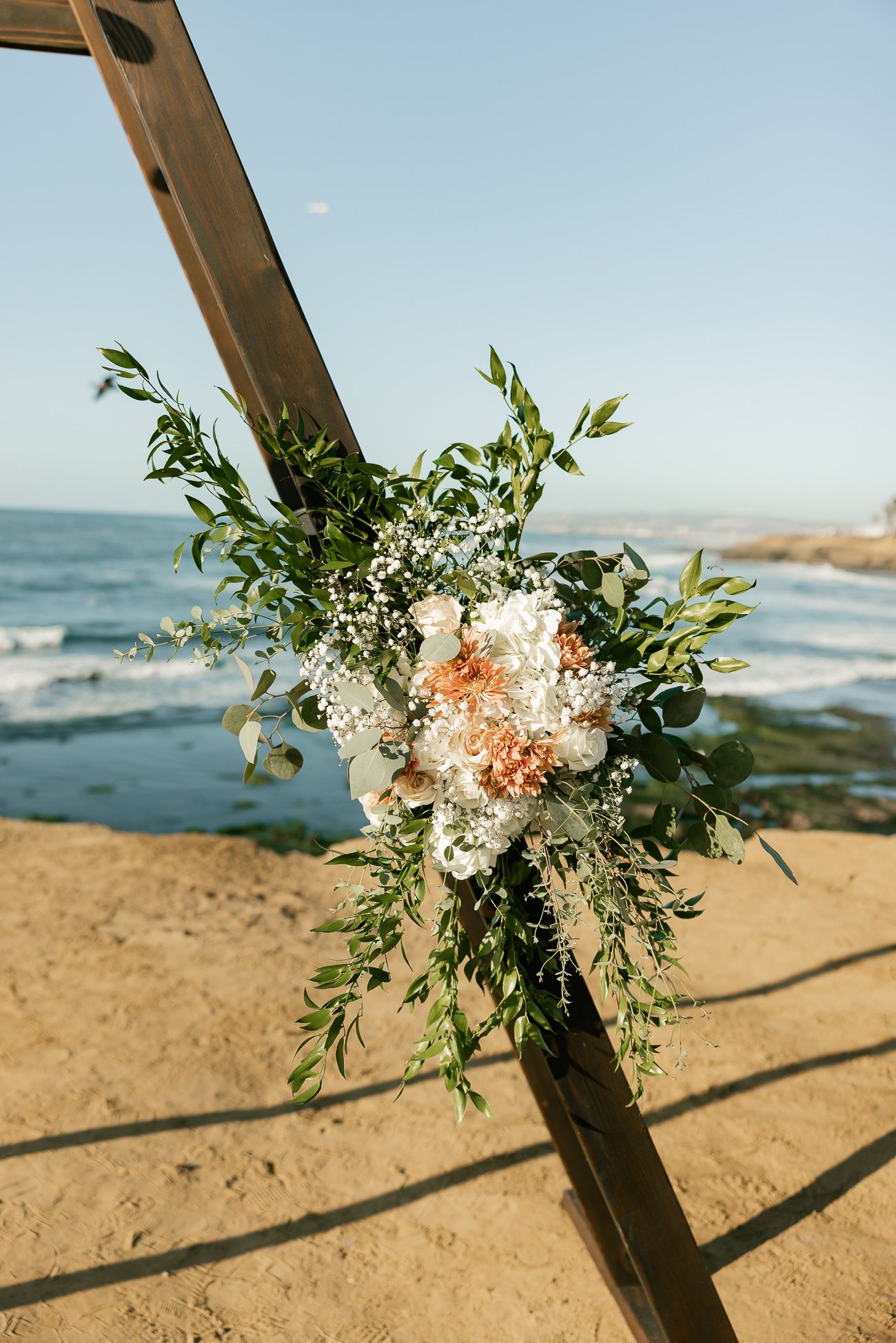 Delicate floral arrangement with white and peach blooms adorns wooden arch on beach with ocean backdrop.