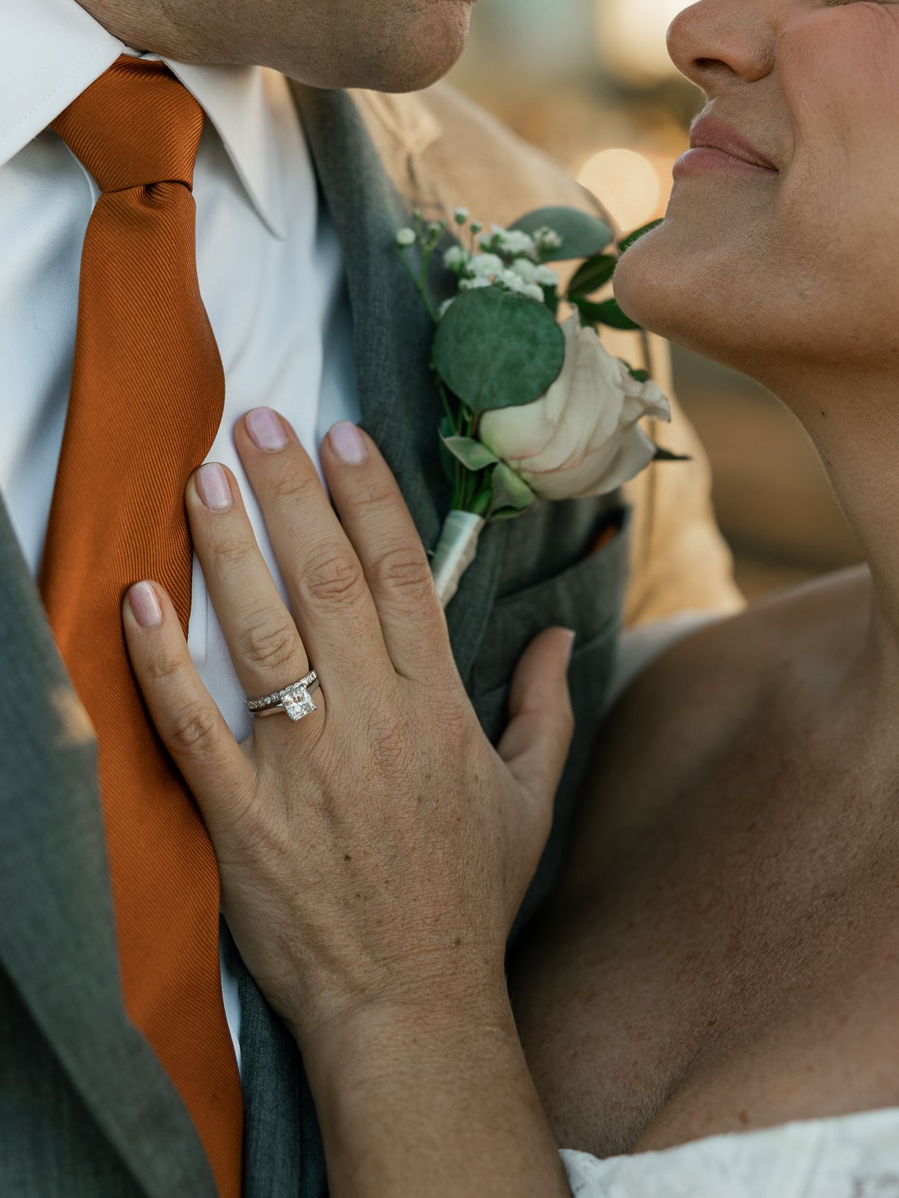 A close-up shot of hands wearing a wedding ring with an orange tie and boutonniere visible.