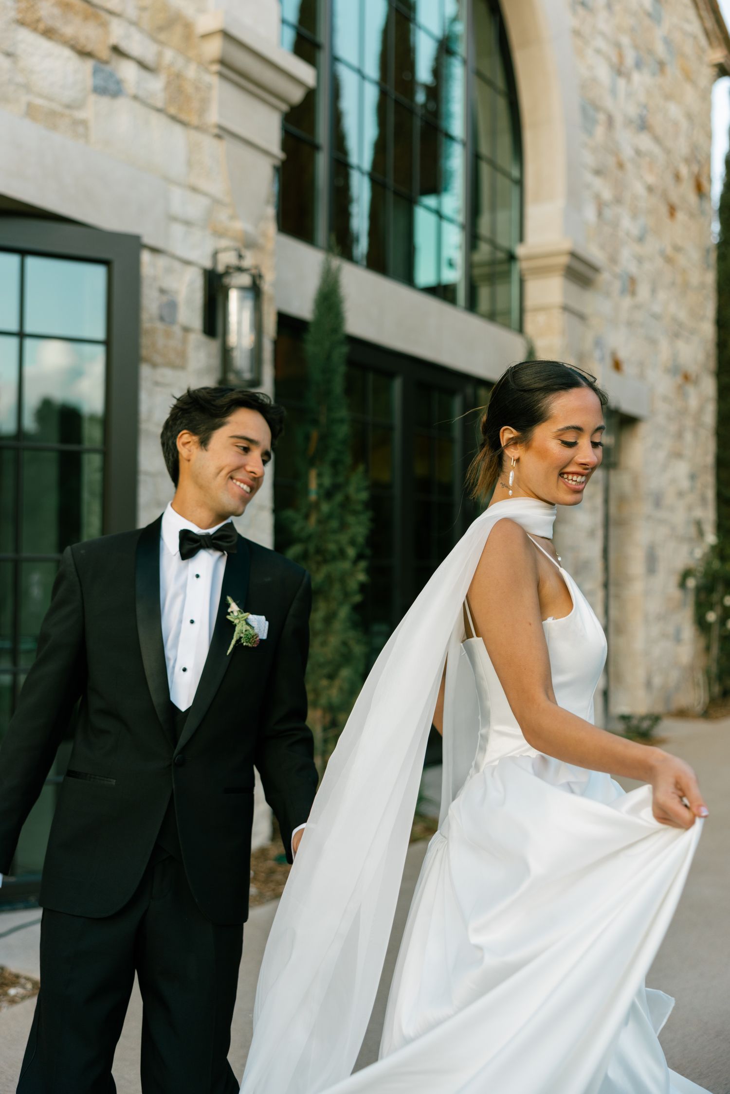 Joyful couple shares a candid moment outside stone building, bride wearing white gown and groom in black tuxedo.