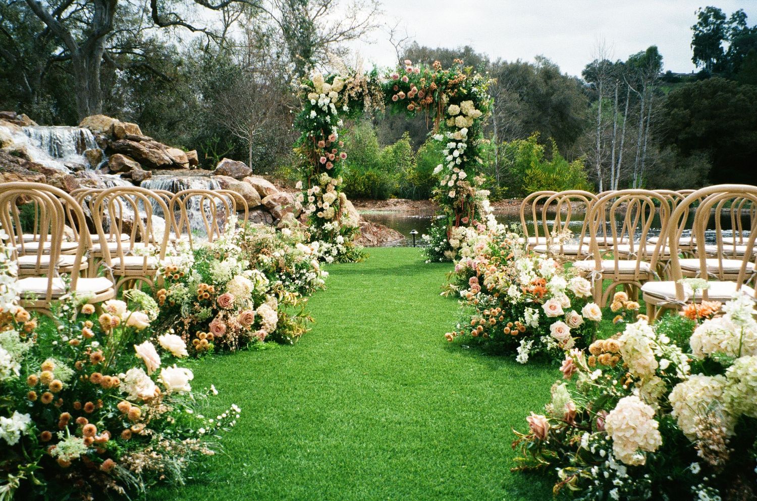Outdoor garden wedding ceremony setup with floral arch and wooden chairs lined along a green grass aisle.