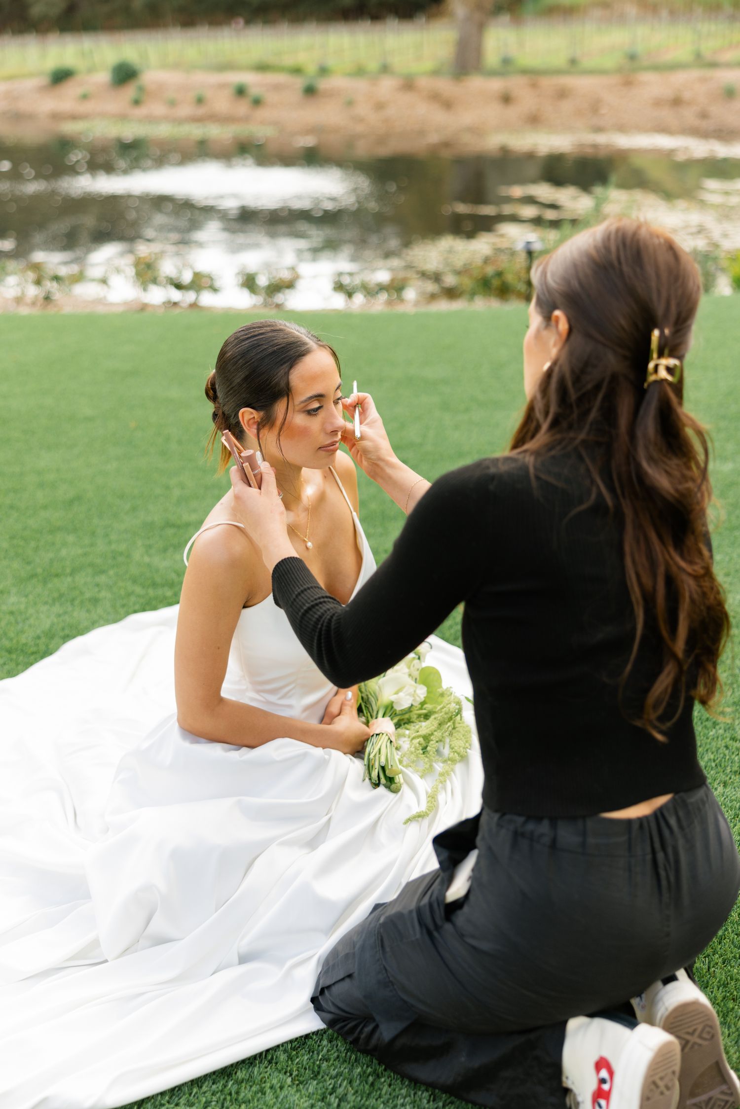 Final wedding makeup preparations taking place on a grassy lawn near a pond.