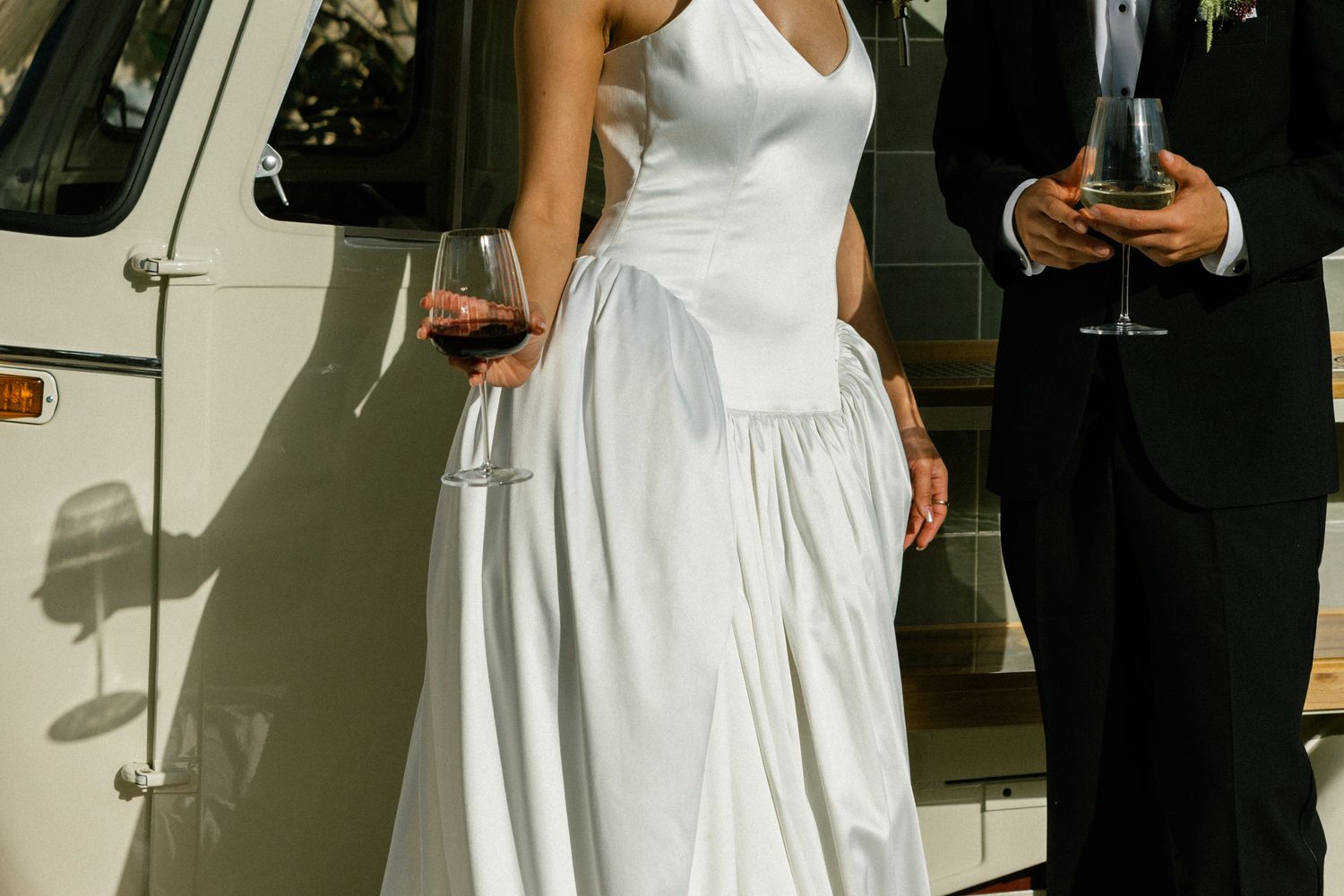A detailed view of a white wedding dress and a person in a dark suit standing beside a classic car outdoors.