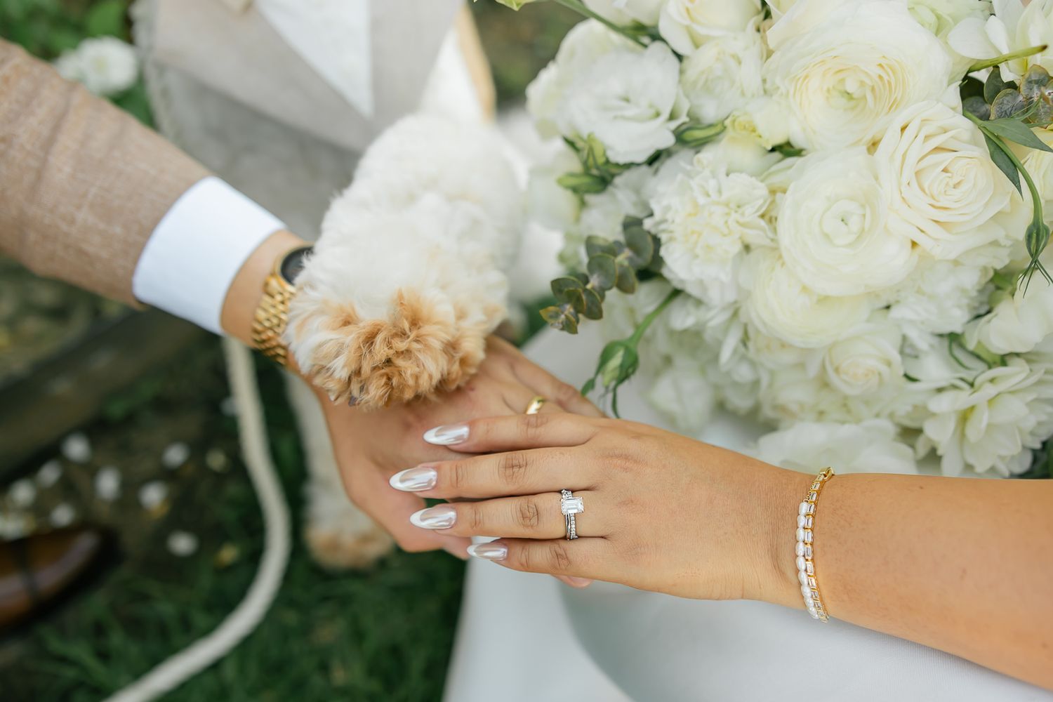 Hands wearing wedding rings with dog paw touch alongside white rose bouquet and bracelet against garden backdrop.