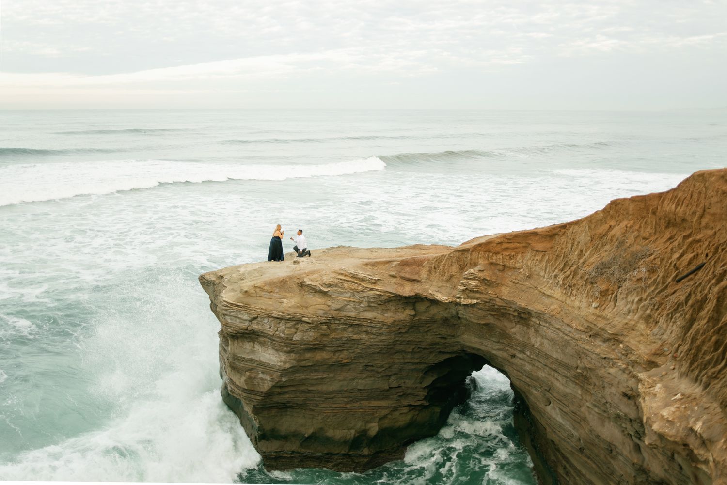 Two people sit on a dramatic natural stone arch formation overlooking crashing ocean waves on a misty coastline.