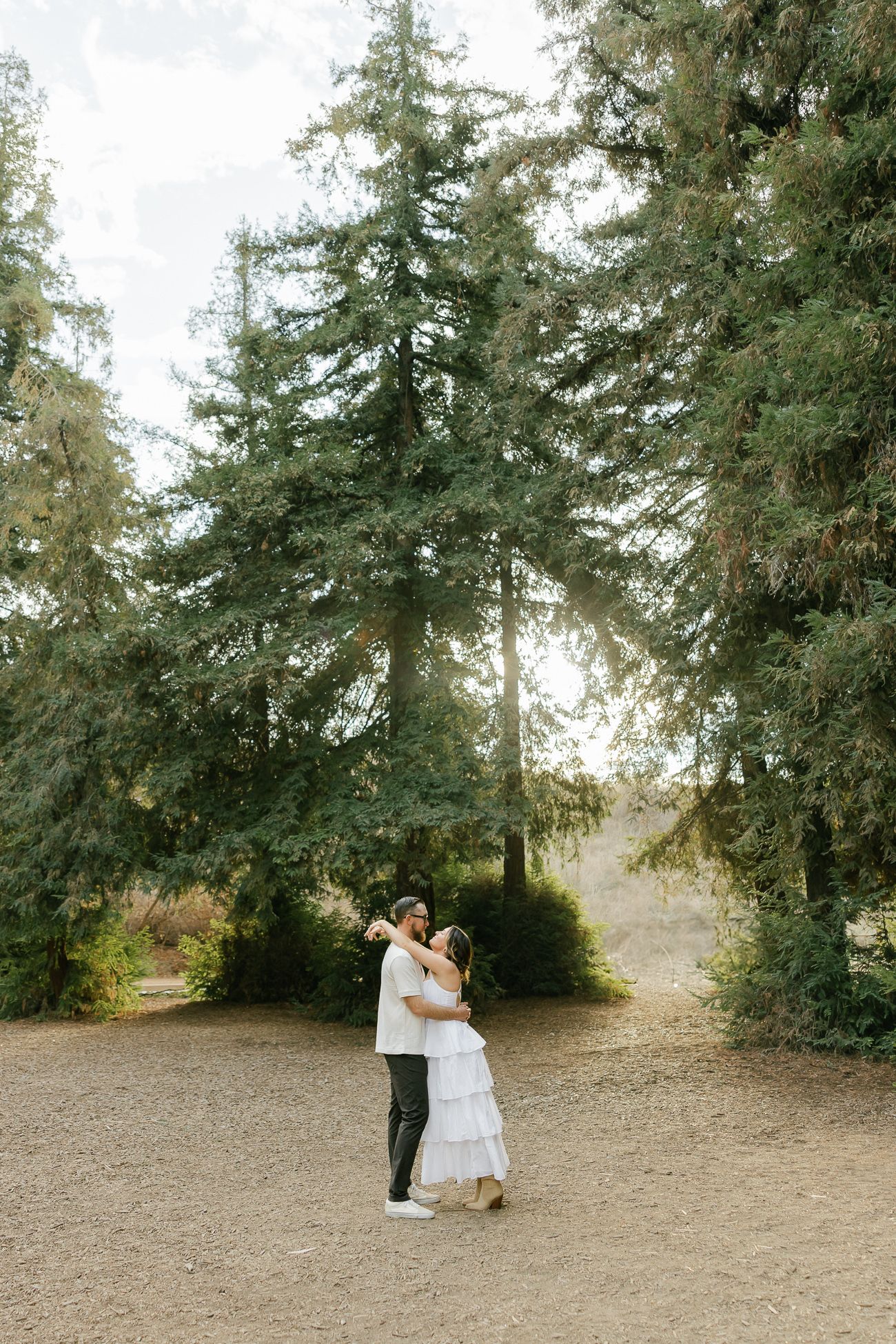 A couple embraces on a dirt path surrounded by tall evergreen trees with sunlight filtering through the branches.