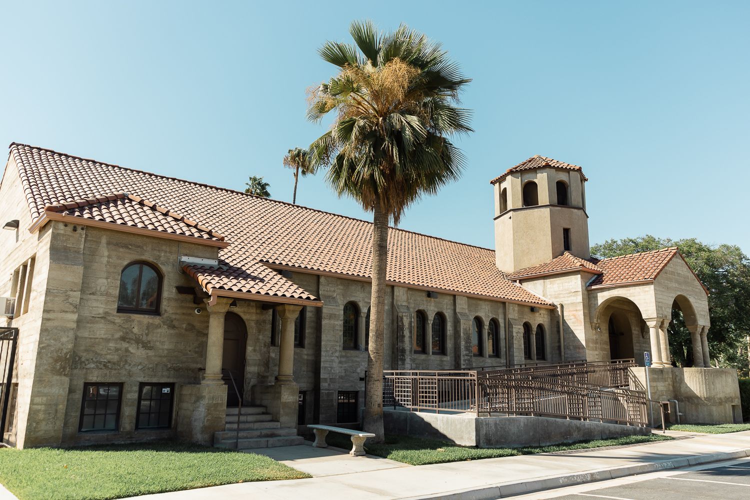 Spanish colonial style church building with red tile roof and bell tower under blue sky with palm tree in California.