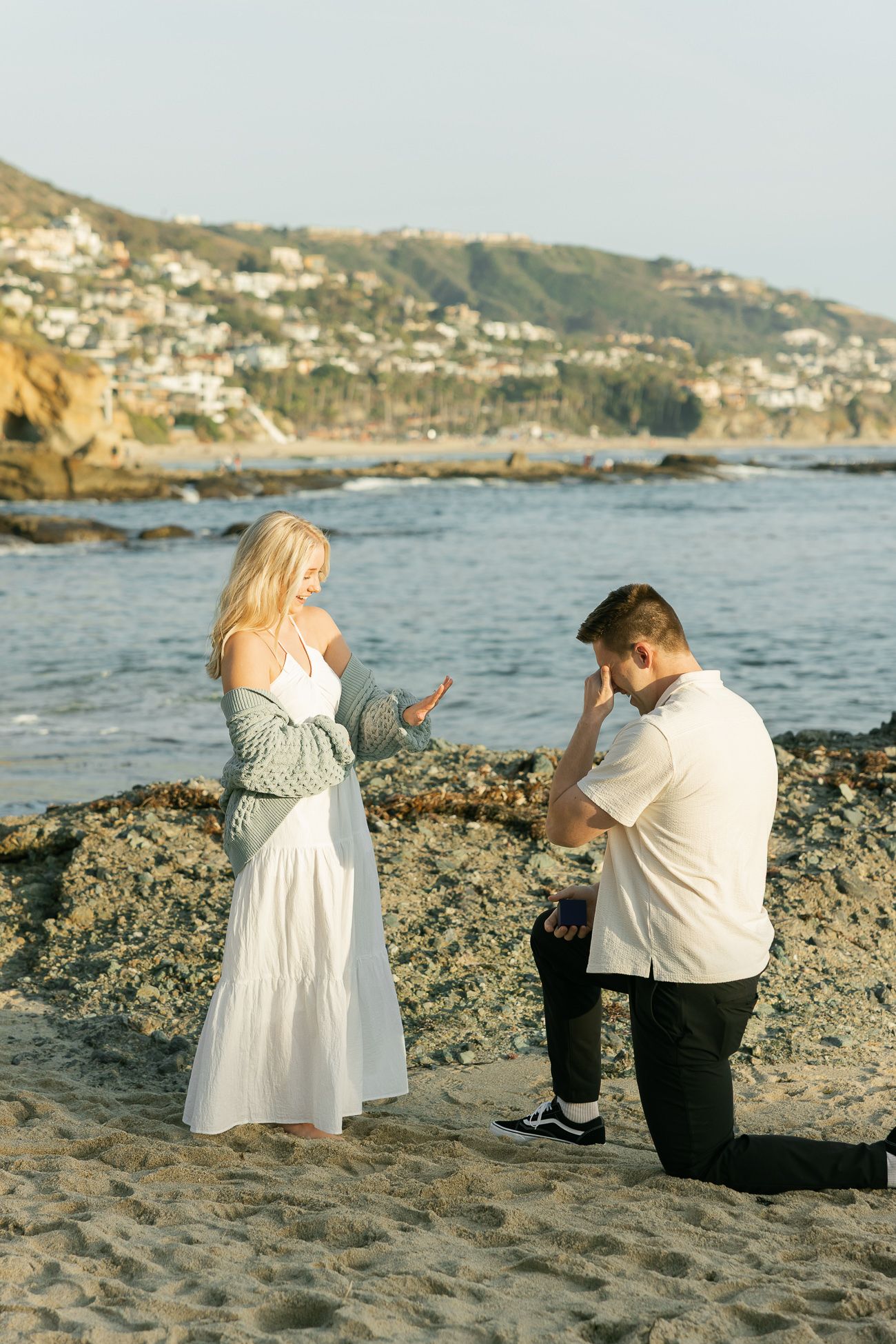 A beachside proposal captured during a sunny afternoon with coastal cliffs in the background.