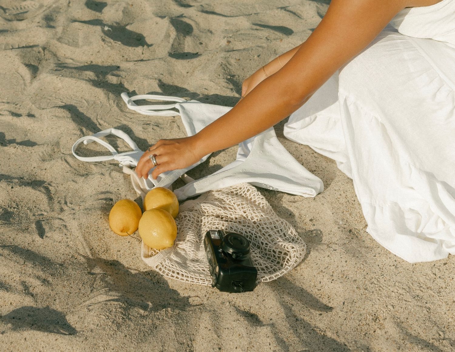 Hands arranging fresh citrus fruits and vegetables in a woven basket on sandy beach surface.