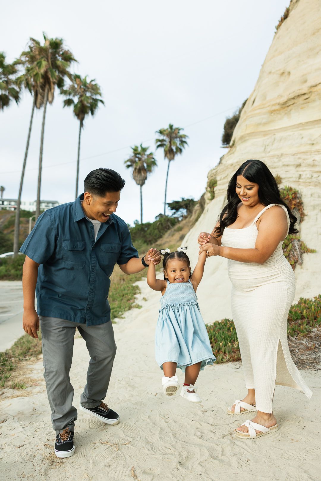 A family of three enjoys a playful moment together on a sandy beach with palm trees and cliffs in the background.
