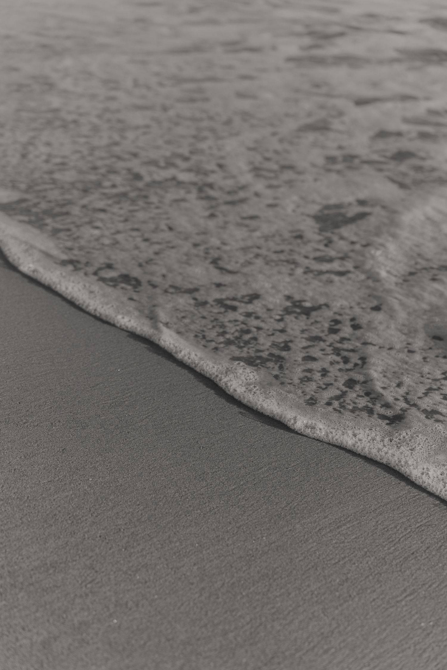 Ocean waves gently roll onto smooth sandy beach in black and white monochrome photography.
