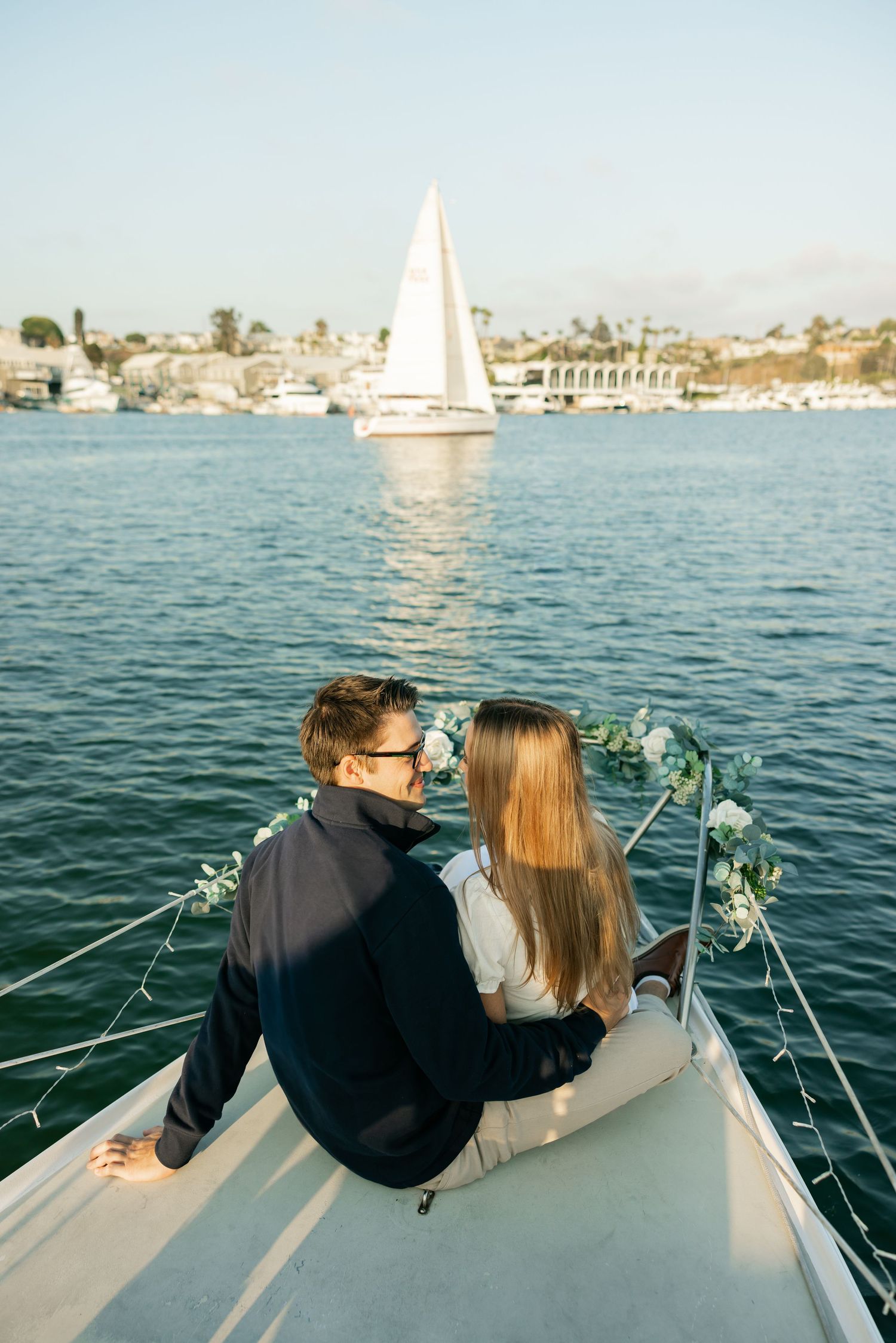 A couple sits together on a sailboat deck while another sailboat glides across the blue harbor waters in the distance.
