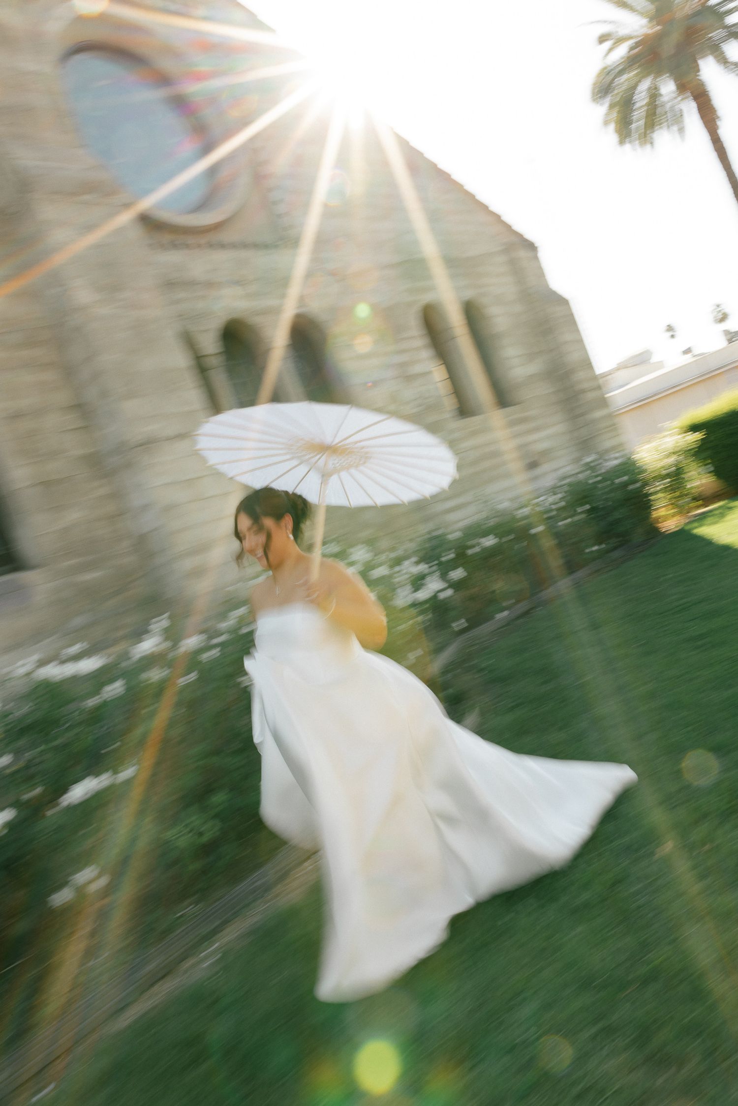 A flowing white dress and parasol create movement against a church backdrop with dramatic sunburst.