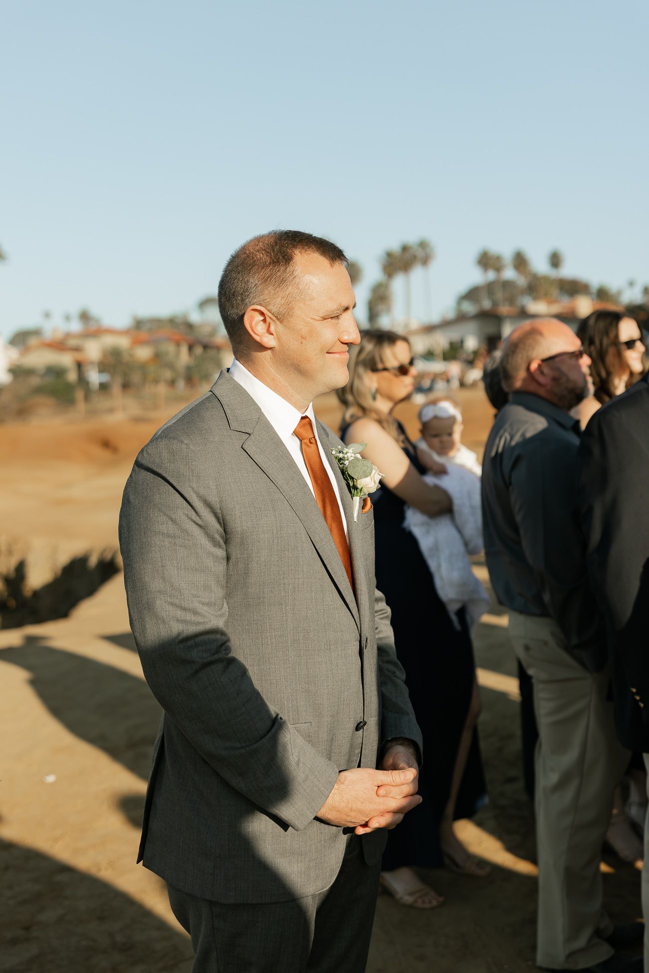 Wedding guest in gray suit with orange tie stands outdoors at scenic coastal ceremony location at sunset.