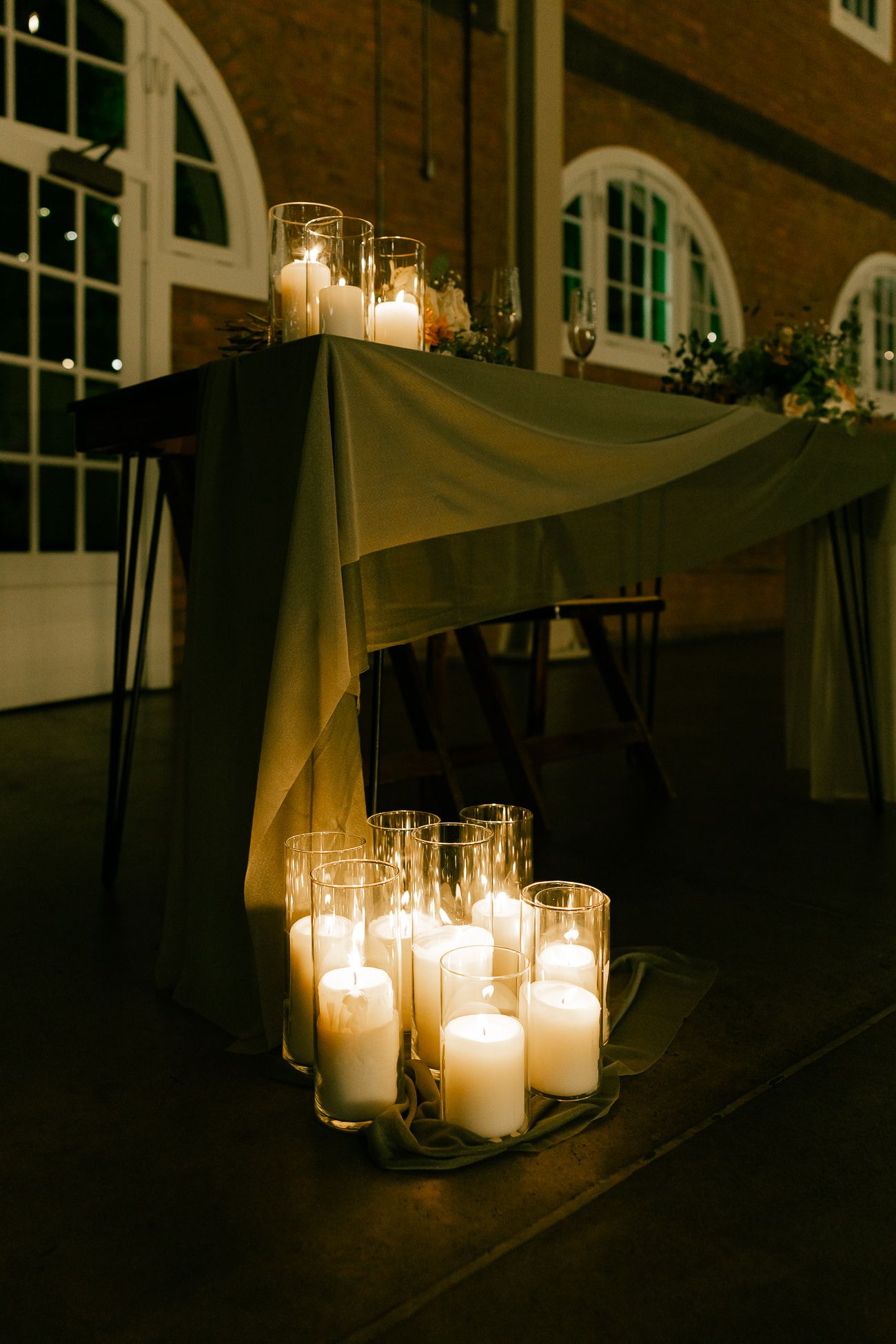 Elegant white candles in glass holders illuminate a draped table setting at an evening reception.