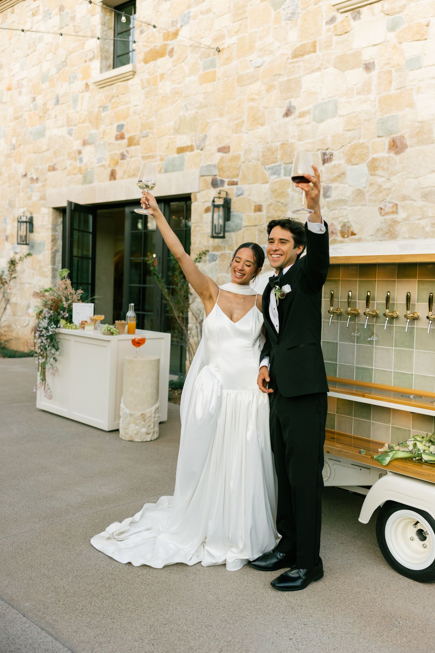 A bride and groom celebrate joyfully outside a stone building with raised arms during their wedding reception.
