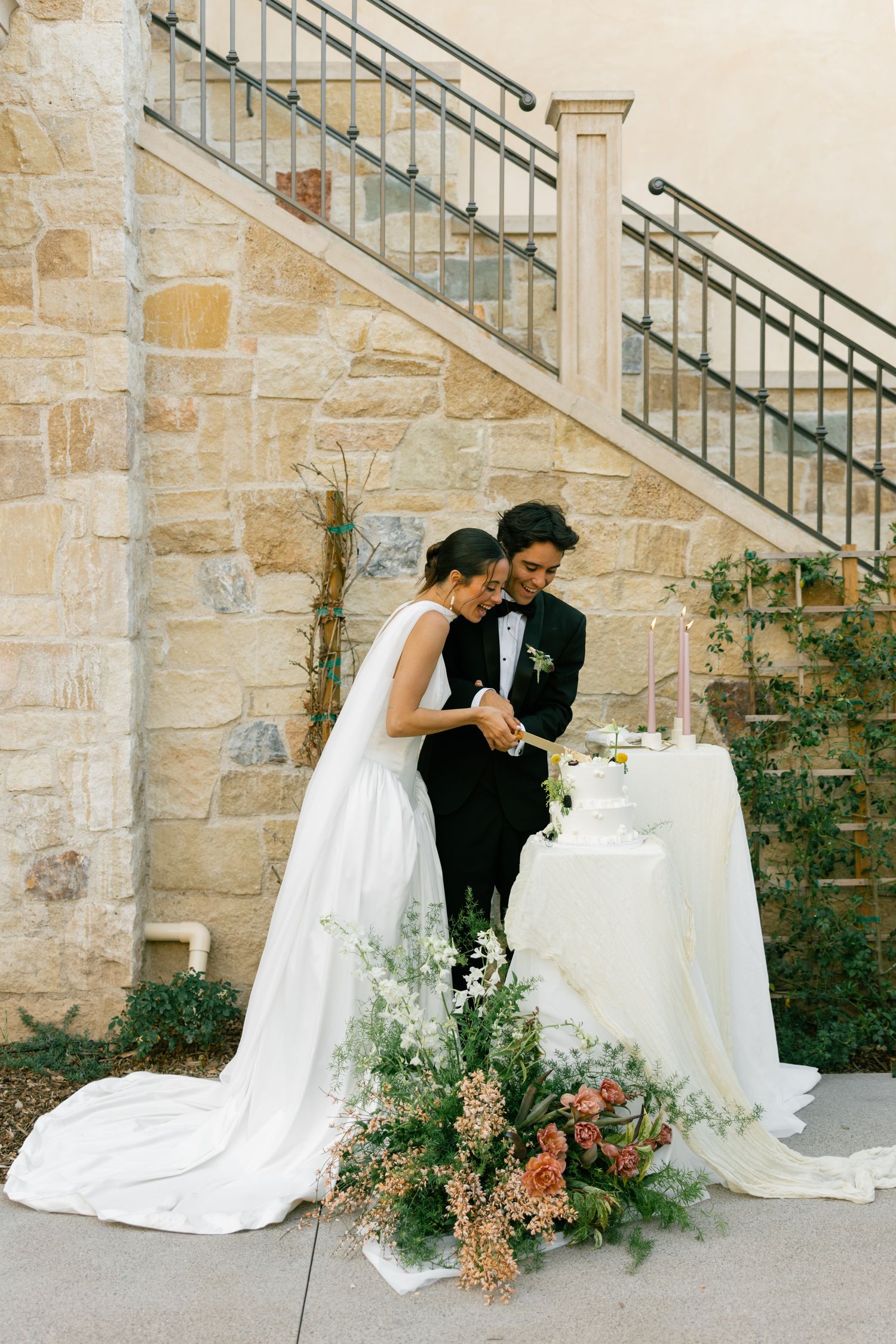 A couple in wedding attire cuts their wedding cake by stone stairs decorated with elegant floral arrangements.